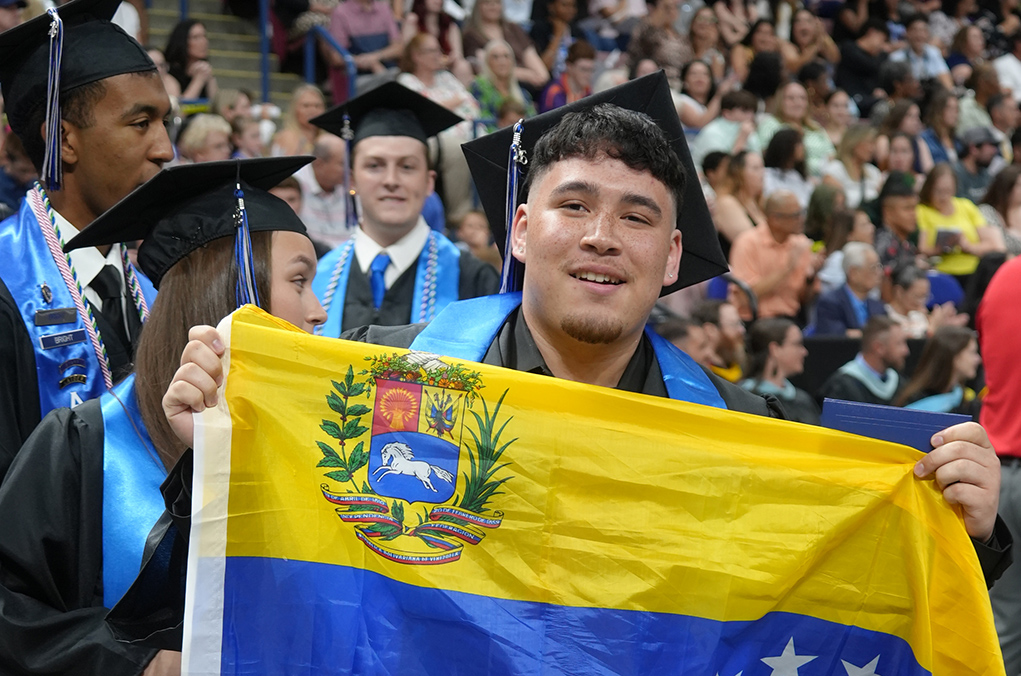 Graduates in caps and gowns, one holding a Venezuelan flag, at an outdoor ceremony.