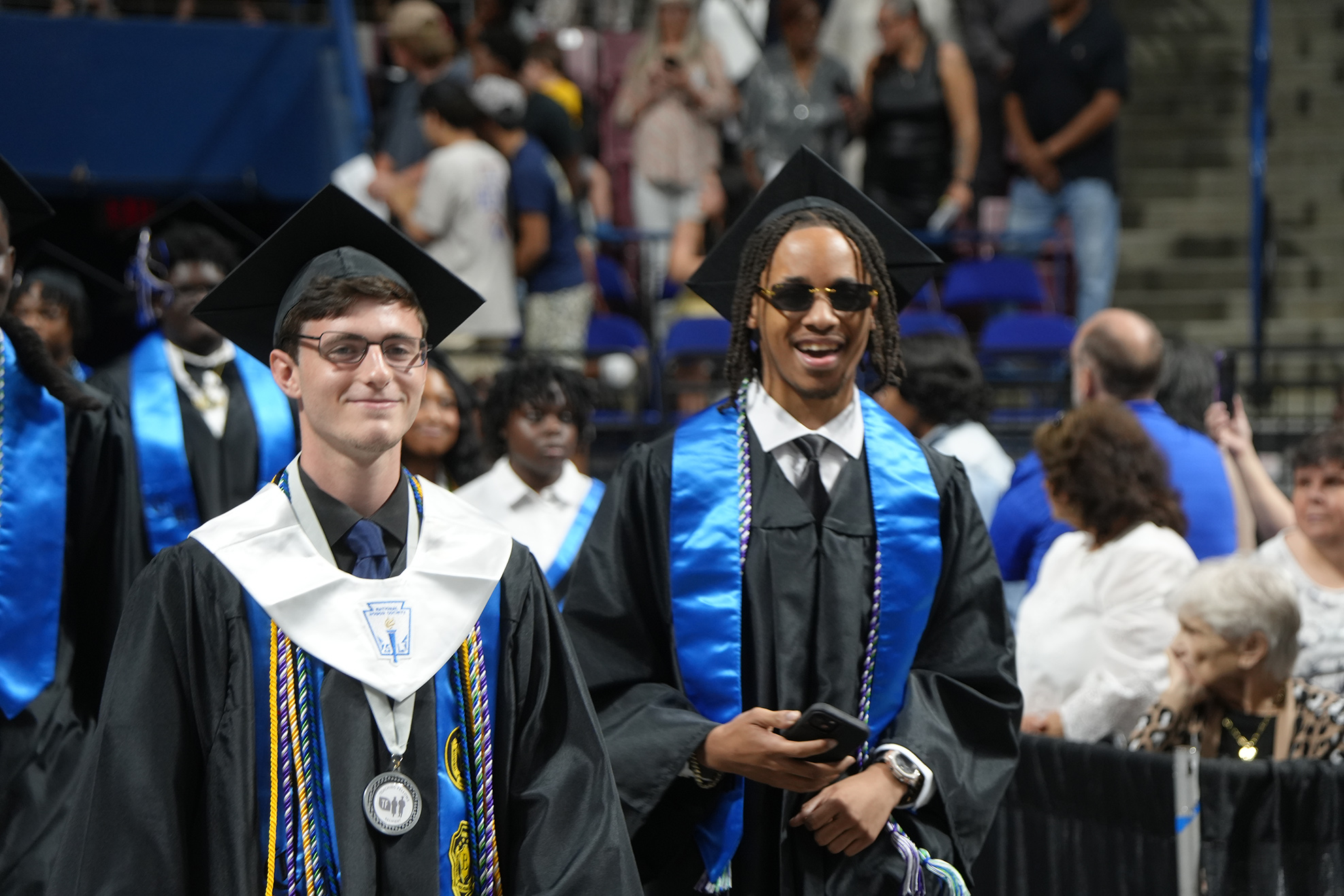 Two men in black academic gowns and mortarboard hats walk in a graduation ceremony. Behind them, a crowd of people stands or sits on bleachers.