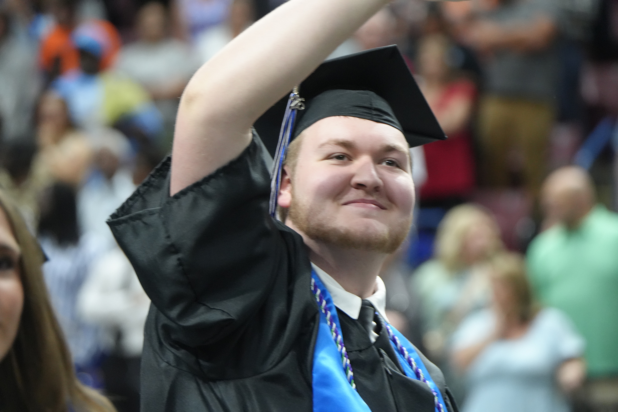Man in black graduation gown raising arm, smiling, surrounded by blurred audience members in the background.