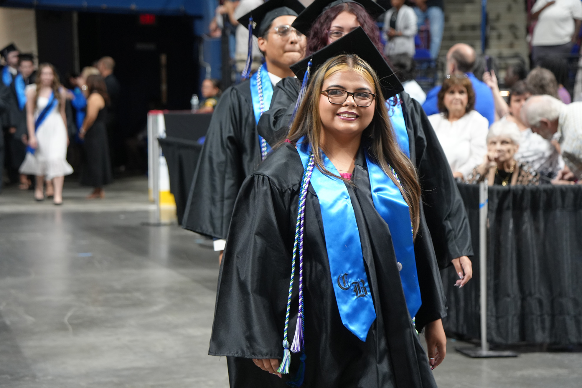 A smiling graduate in a black gown with a blue sash and cap walks with others. Behind, spectators stand and watch.