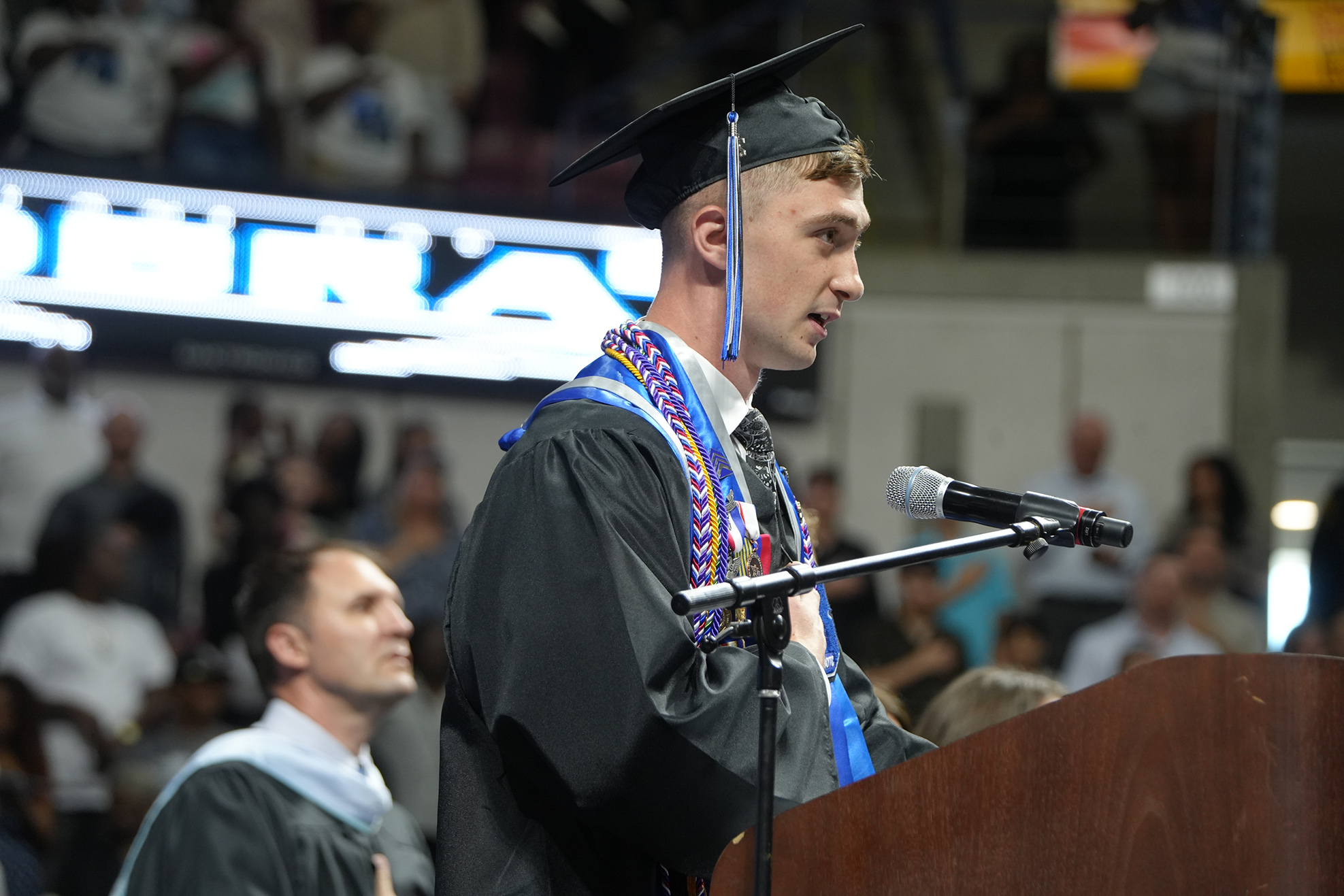 A male speaker at a graduation ceremony speaks into a microphone, wearing a graduation gown and cap. Behind him, a crowd of people is seated in the audience.