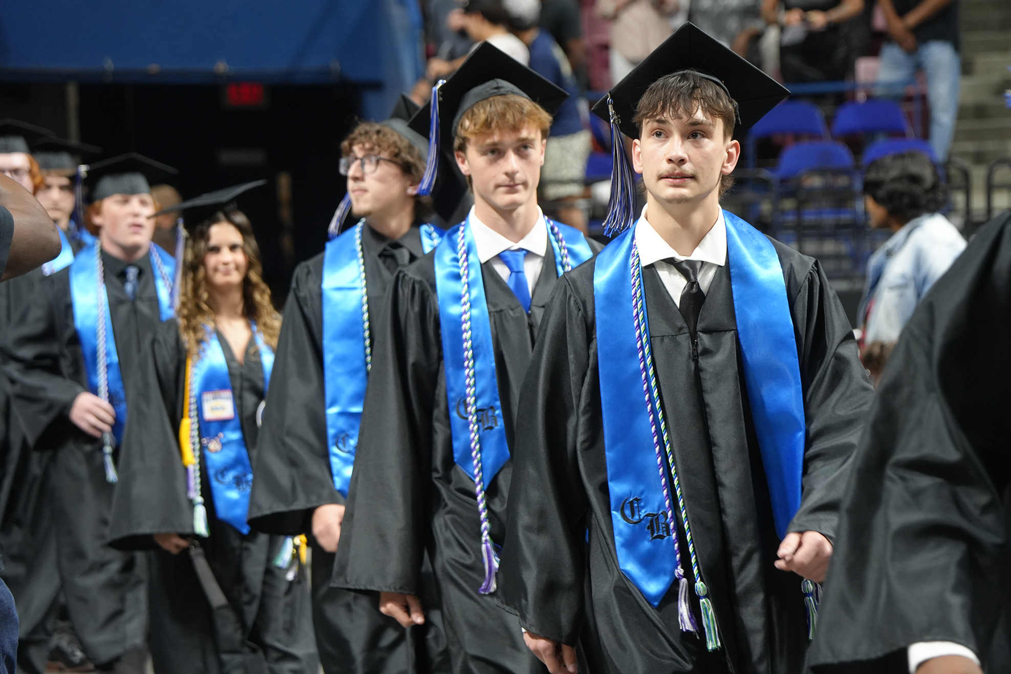 A line of young men and women in black gowns and blue stoles walk across a stage in a stadium.