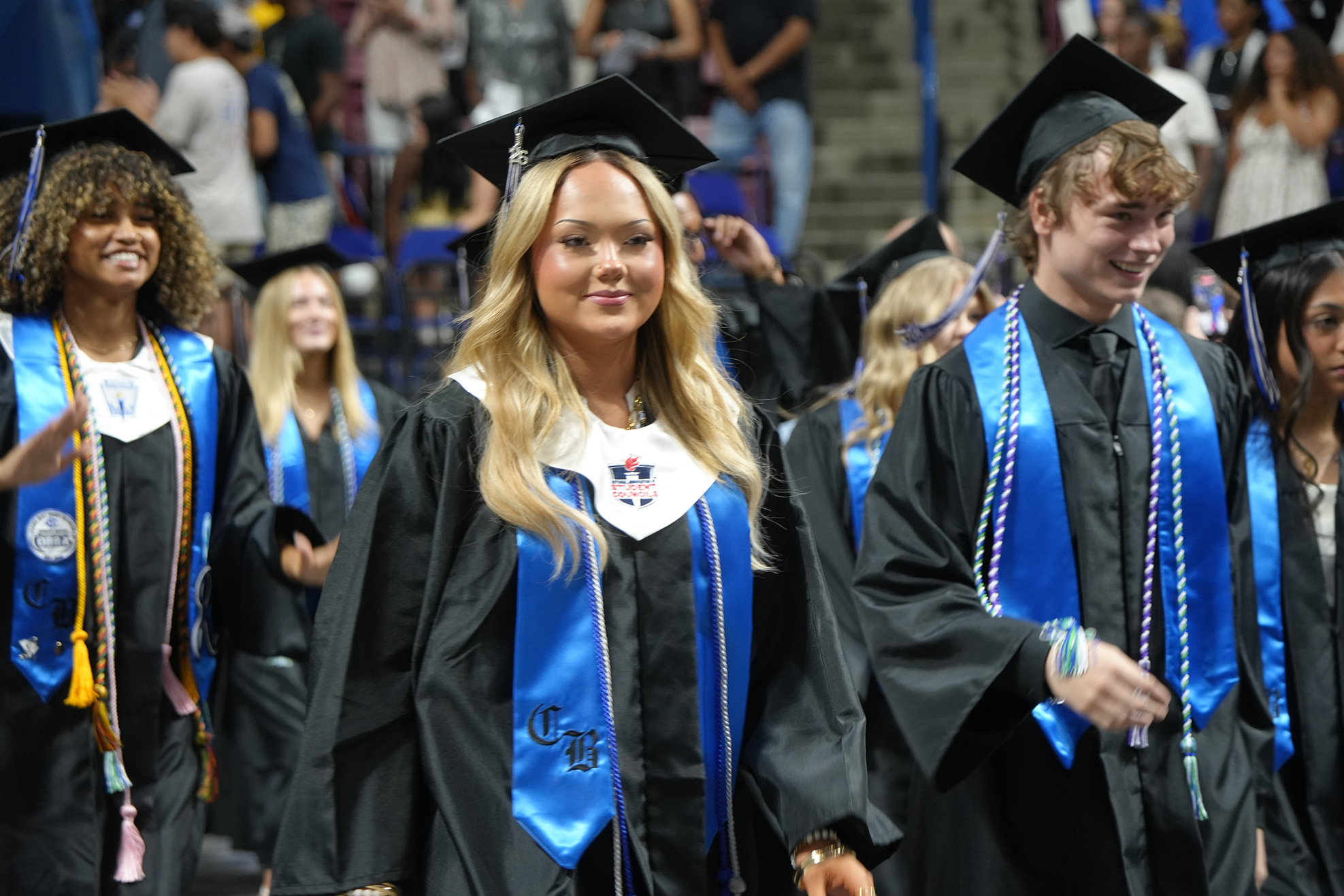 Graduates in black gowns and blue stoles walk in a ceremony. Behind them, students in similar attire walk past.