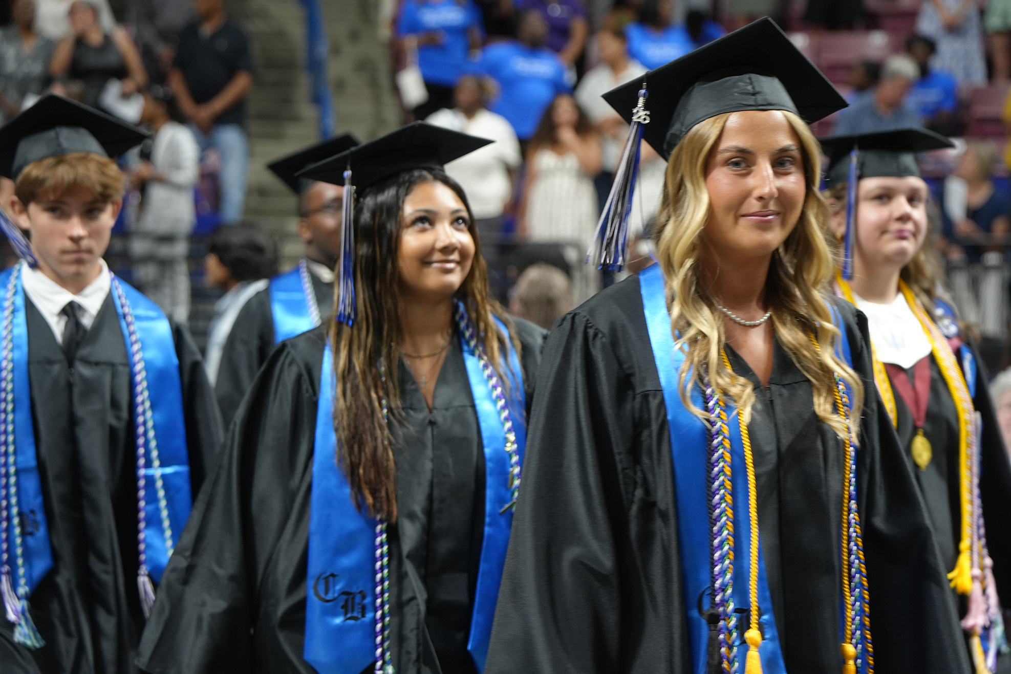 Graduates in black gowns and blue stoles, wearing caps, walk in a line at a ceremony.