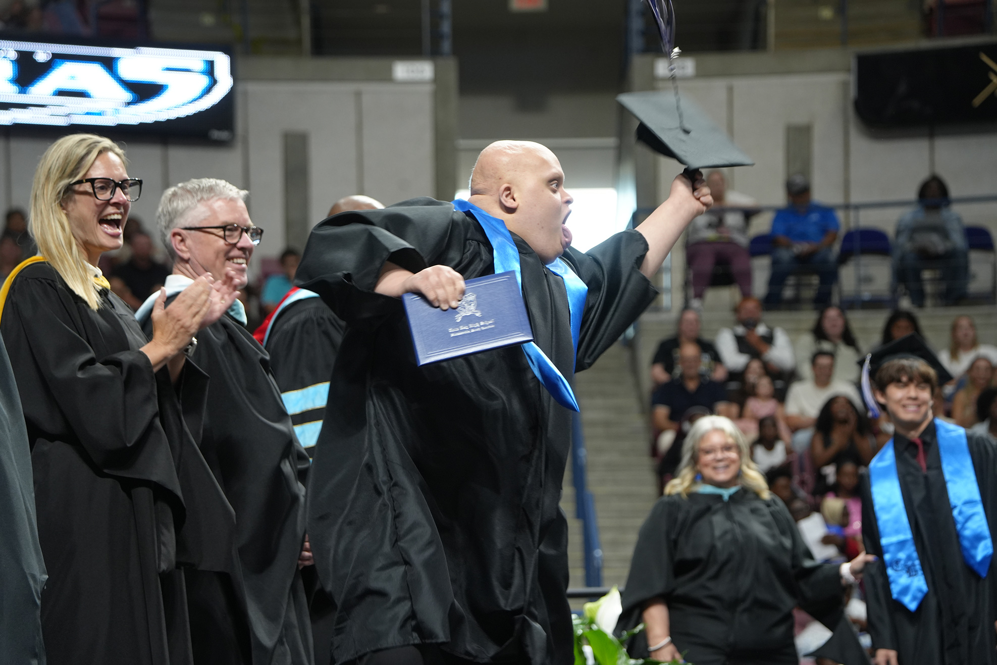 Graduates in black gowns at a ceremony, one man removing his cap and holding it up, others clapping.