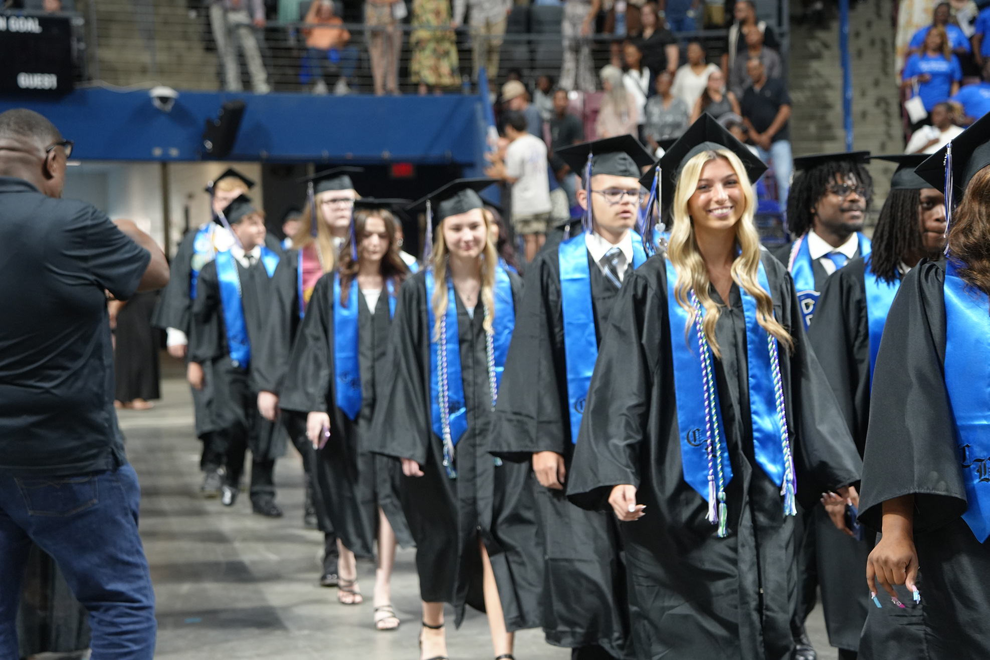 Graduates in black gowns and blue stoles walk down the aisle at a ceremony; some wear caps.