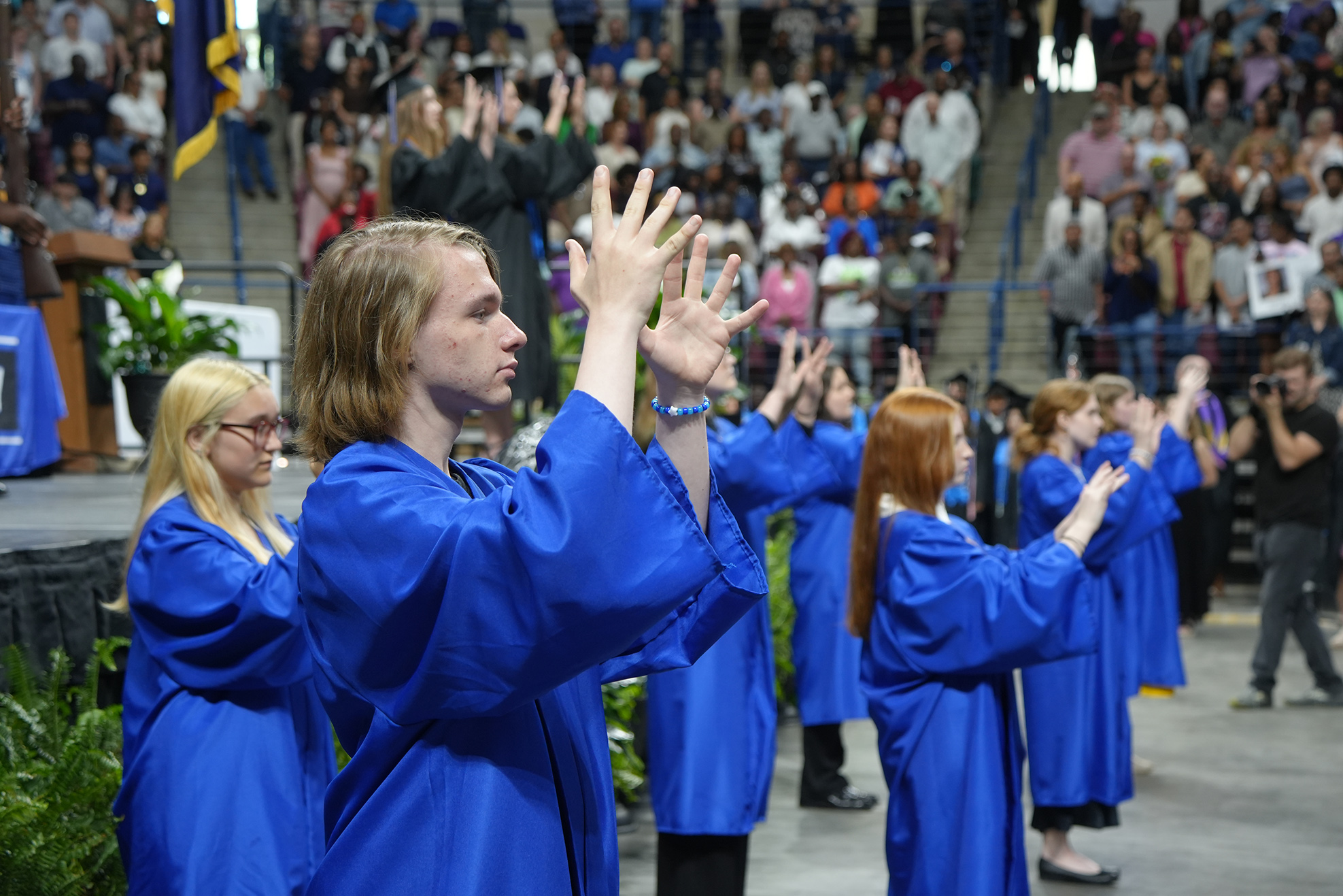 People in blue gowns face an audience at an event; some raise their hands in applause.