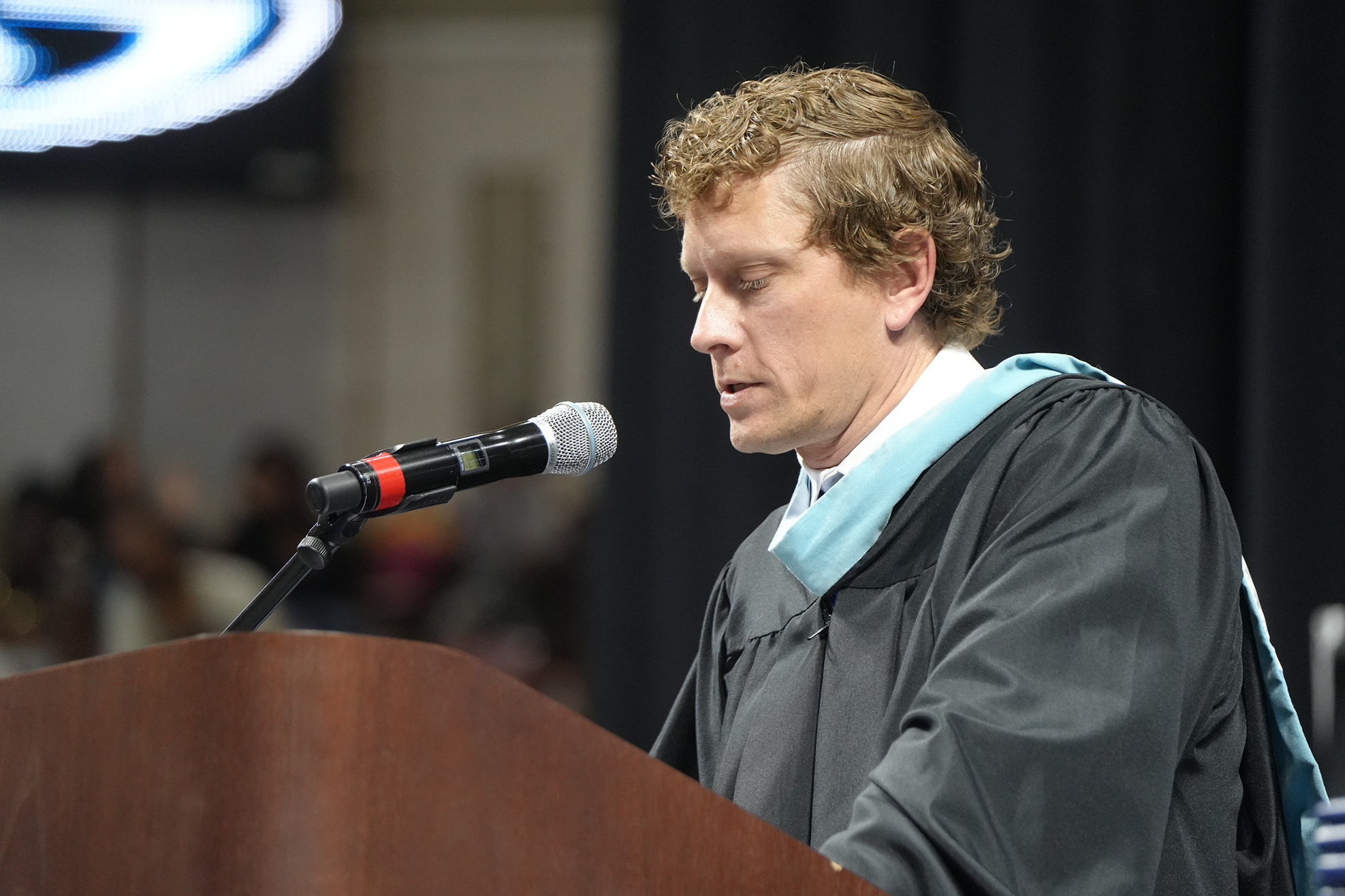 A person in a graduation gown speaks at a podium with a microphone. Behind them, people sit in a room.