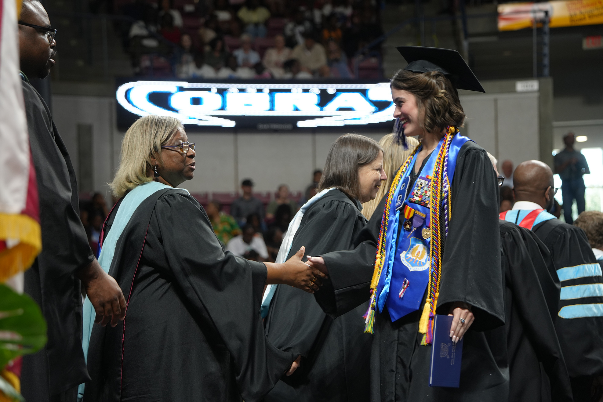A person in a graduation gown shakes hands with another person, both wearing glasses. Background includes people in gowns and a sign.