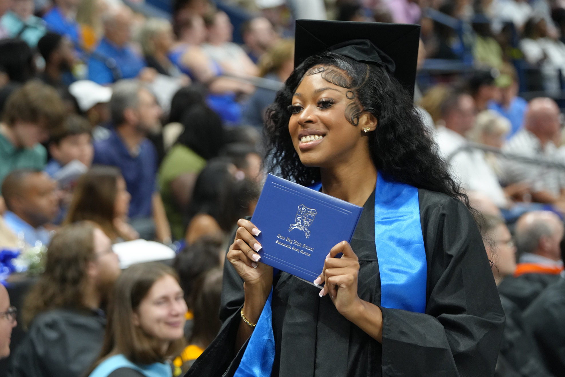 A woman in a black and blue graduation gown holds a blue folder, smiling for a photo at an event.