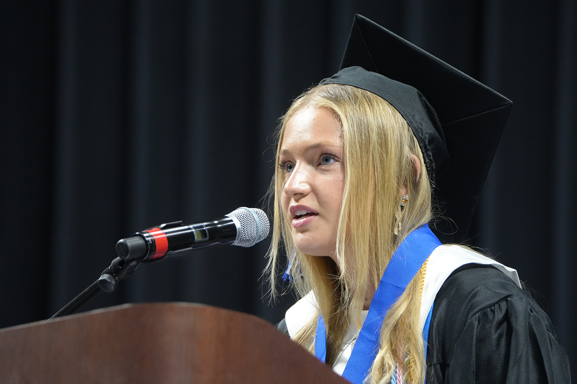 A woman in a black graduation gown and cap, with long blonde hair, stands at a podium speaking into a microphone.