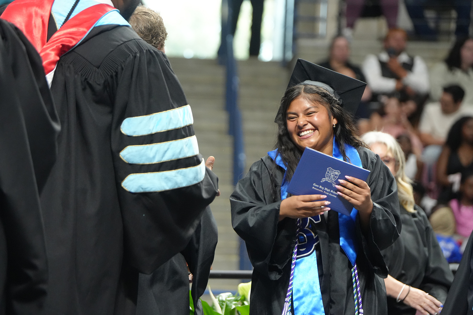 A woman in a blue gown holds a blue folder while smiling, with people in academic gowns around.