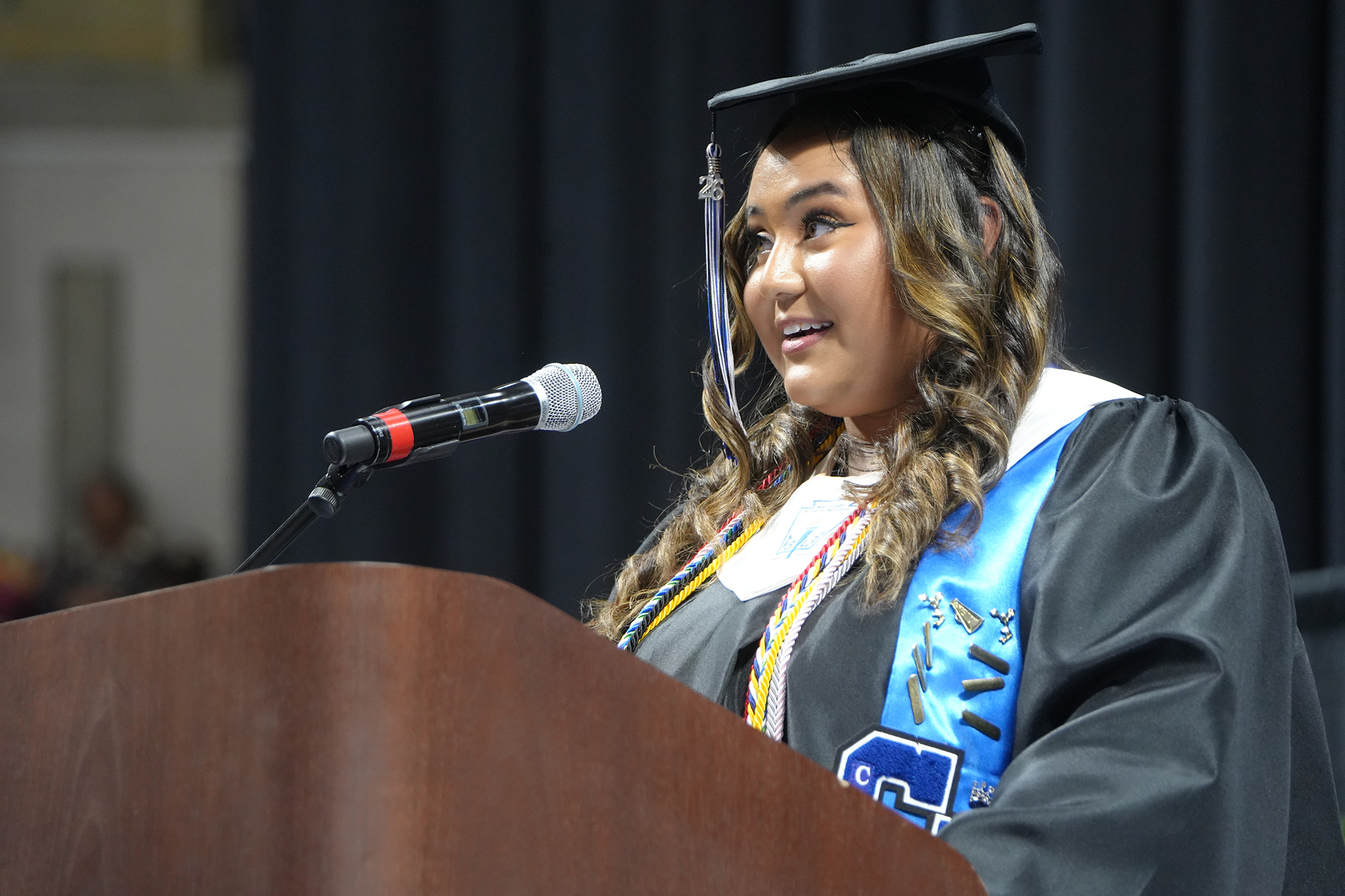 Woman in academic gown and cap standing at podium with microphone, looking up, smiling.