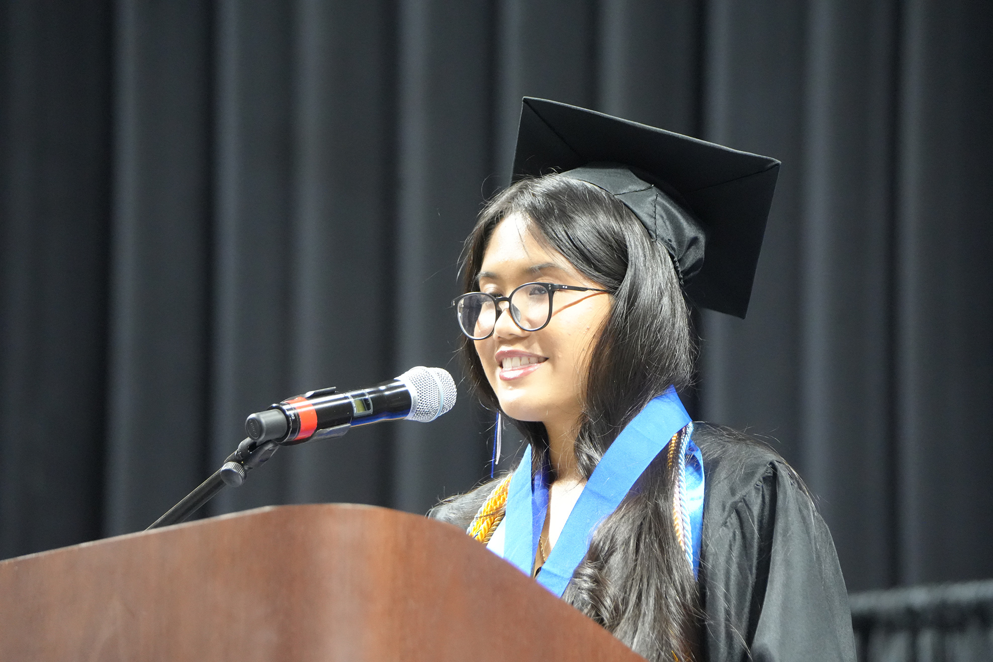 Woman in cap and gown at a podium, speaking into a microphone. Black curtain in background.