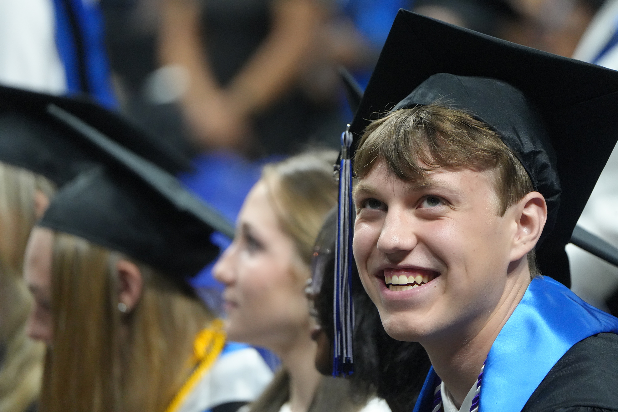 Smiling graduate in a black cap and gown, with a blue stole, surrounded by others in similar attire.