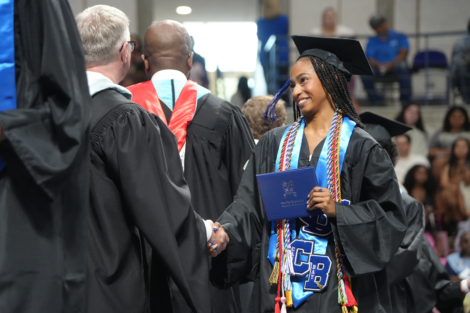 A graduate holds her blue diploma and shakes hands with a faculty member at a commencement ceremony.