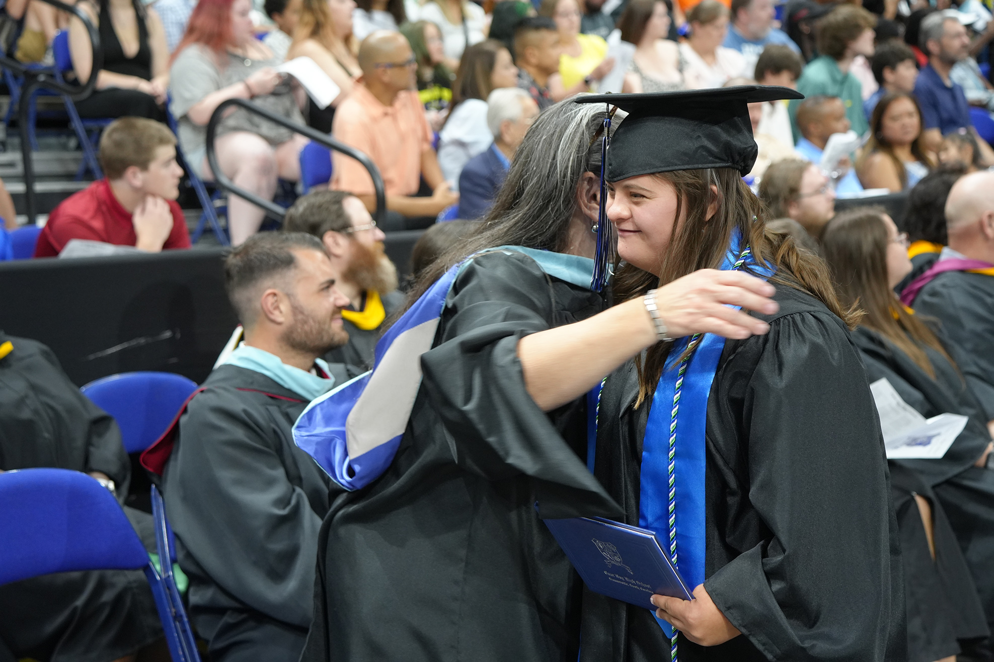 Graduates in gowns and caps celebrate at an indoor event, with audience members seated in the background.