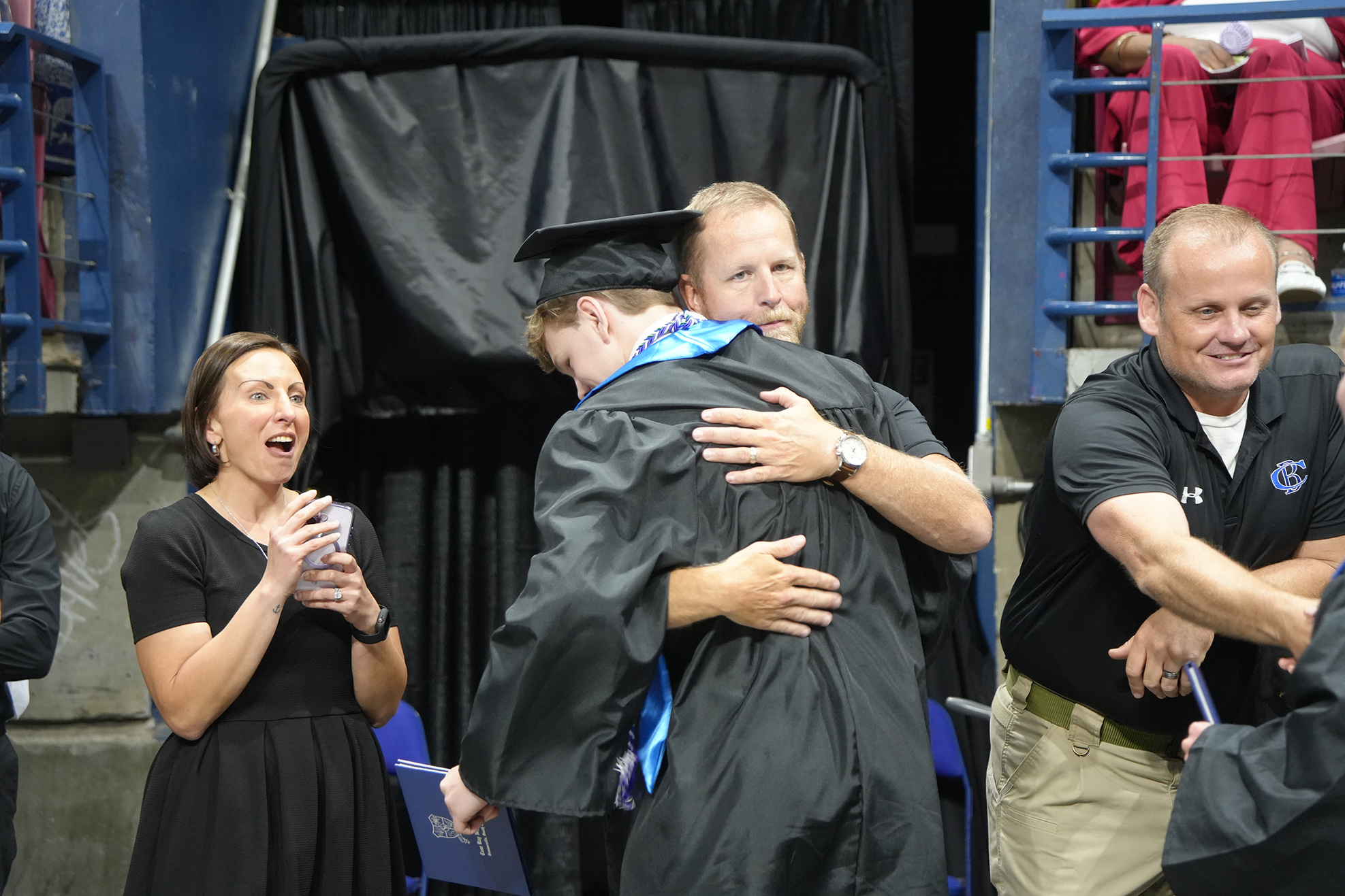 A graduate hugs a man in a black gown, smiling. Two women and a man in a black shirt watch from behind.