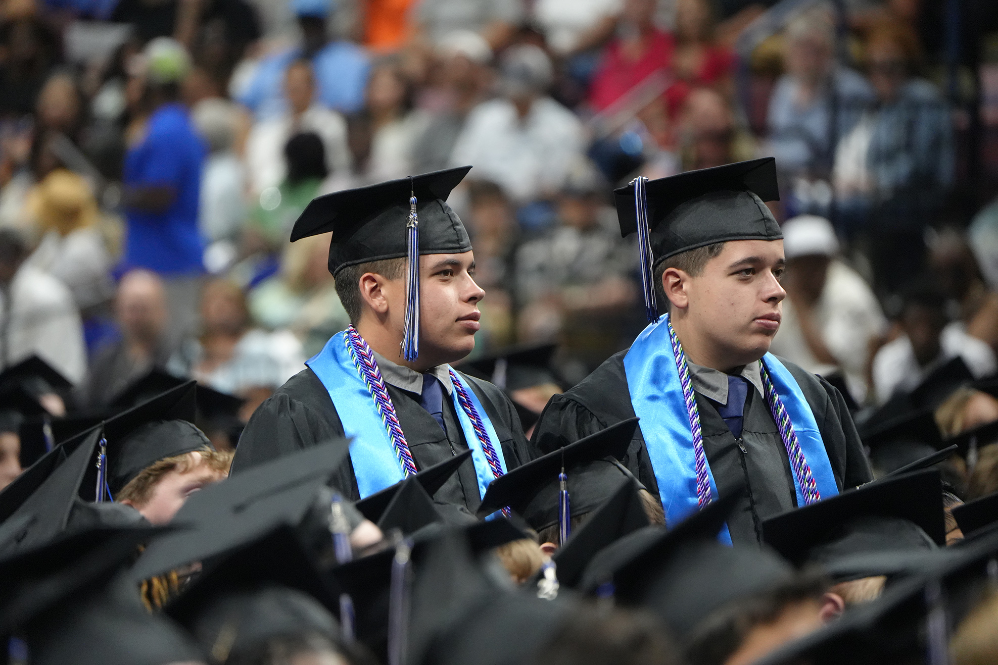 Two men in graduation gowns and caps sit in a large audience of people at a graduation ceremony.