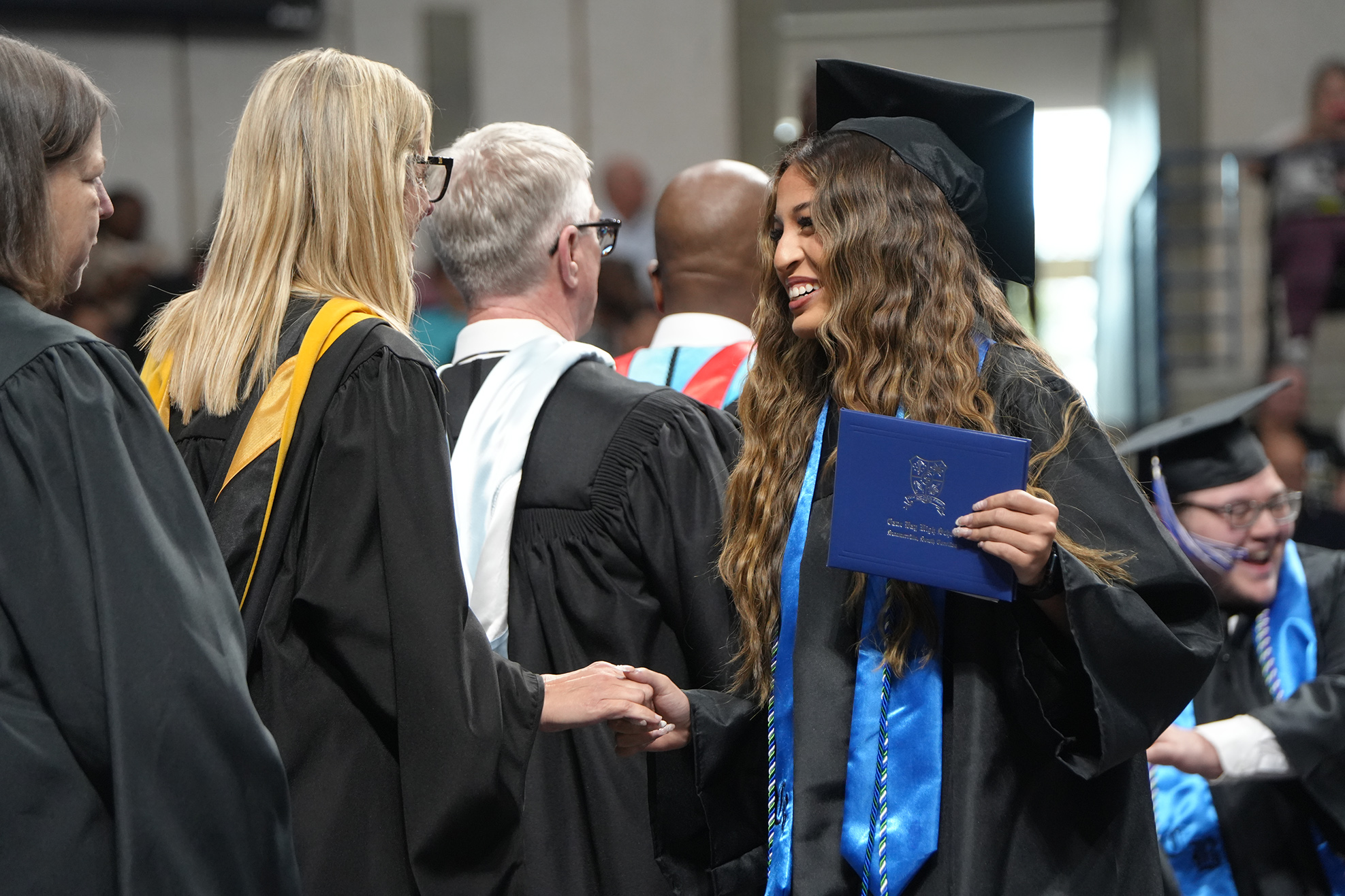 People in caps and gowns stand in line at a graduation. A graduate smiles and shakes hands with faculty.
