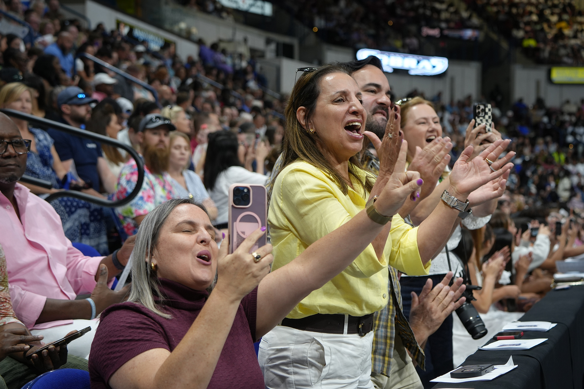 Spectators cheering, raising hands, and holding phones in a sports stadium. Tables with papers and a microphone in front.