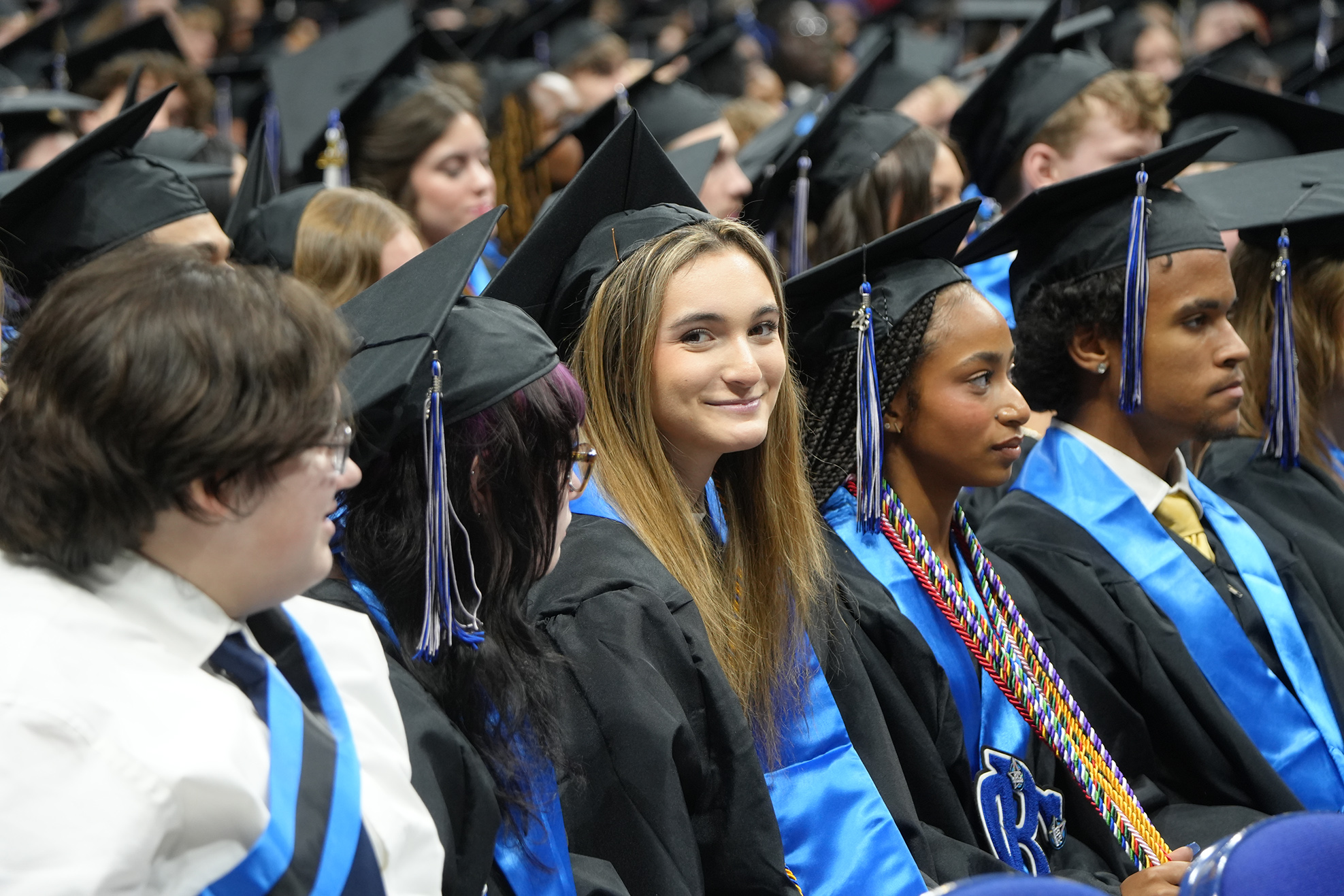 Several students wear graduation gowns and caps. They sit in rows. One person wears a white shirt.