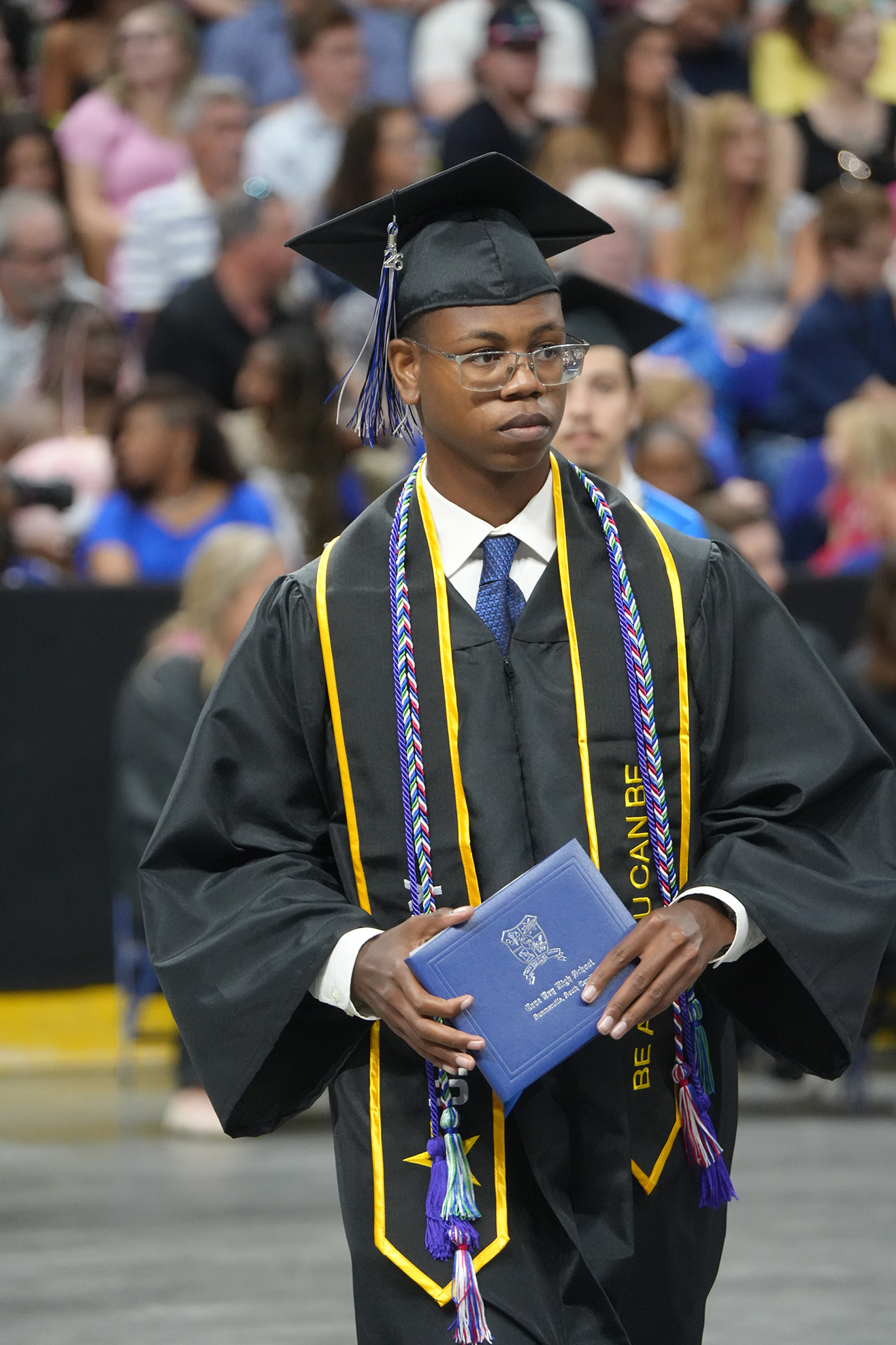 A graduate holds a blue diploma and stands in front of an audience in a black gown and cap.