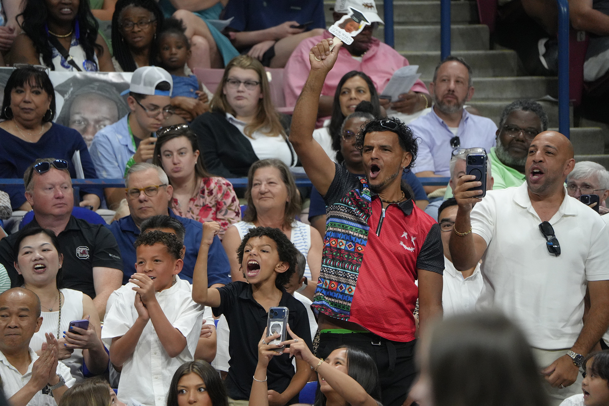 Audience at a sports event, many holding phones and cheering, with a person standing in the center with raised arms.