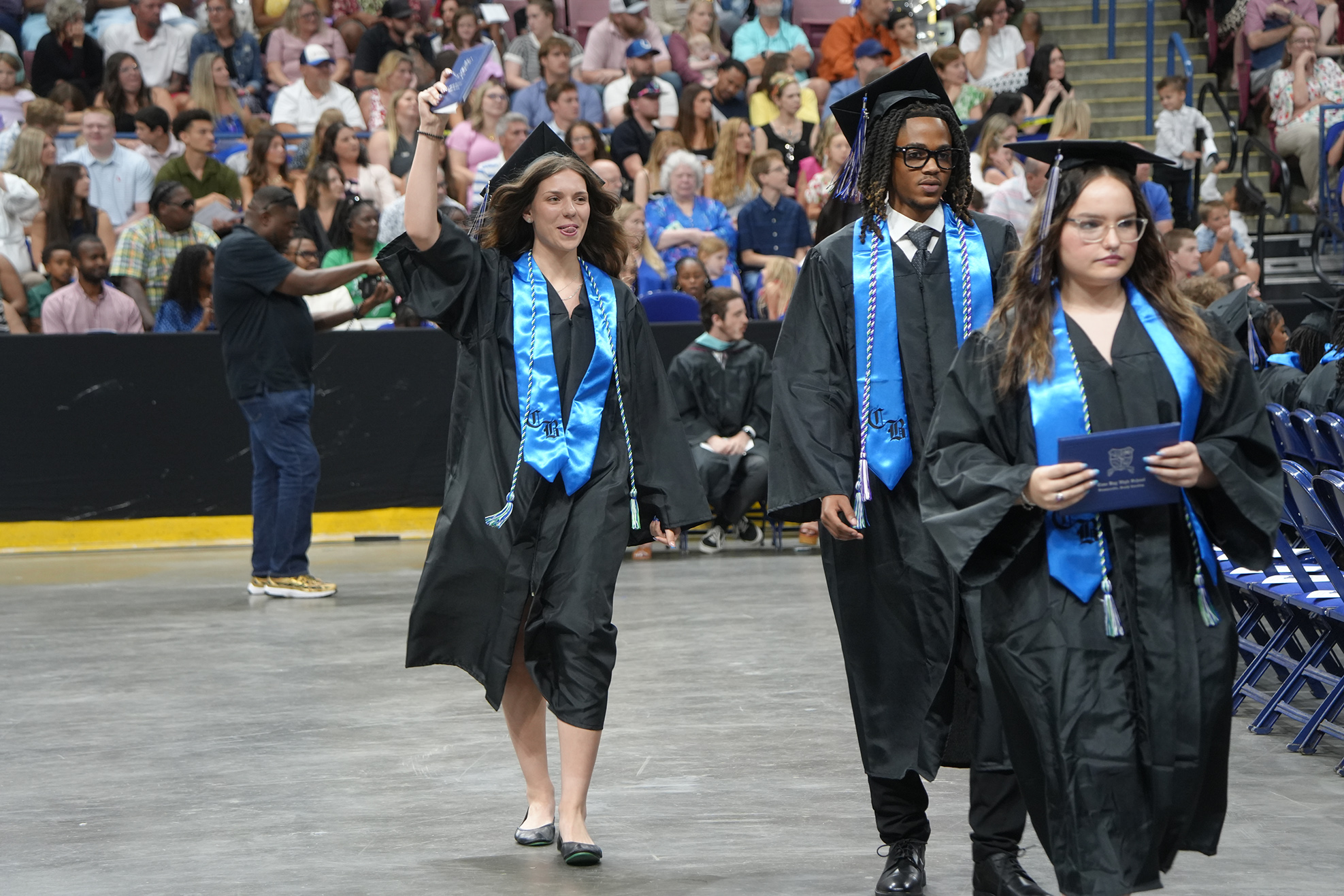 Three people in black gowns and mortarboard hats walk on stage; one raises a diploma. Behind them, a large crowd watches from bleachers.
