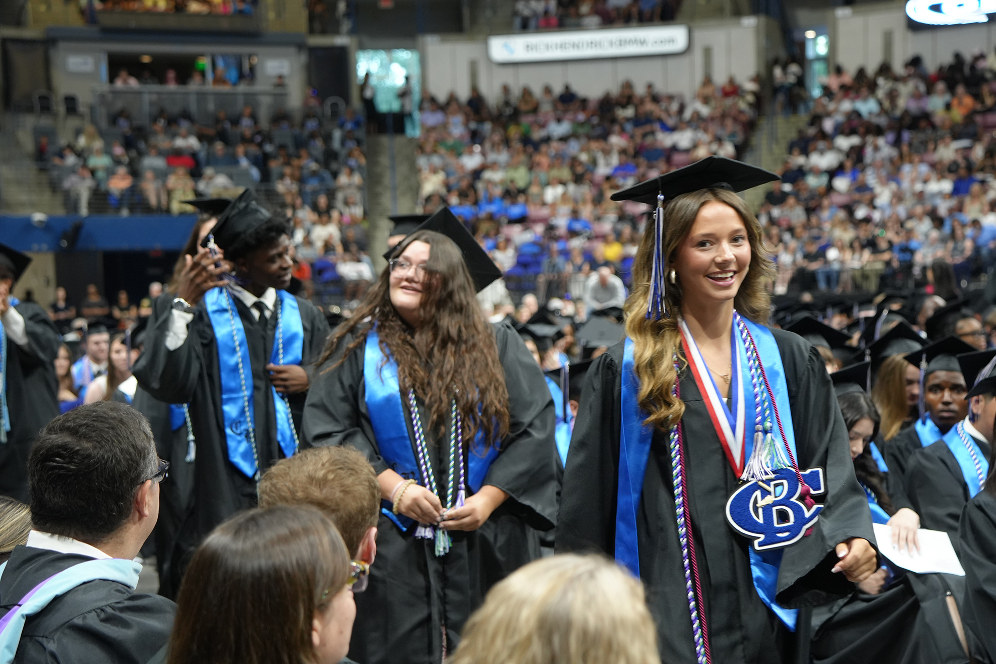 Graduates in blue gowns and caps are standing in an auditorium, smiling and posing for a photo.