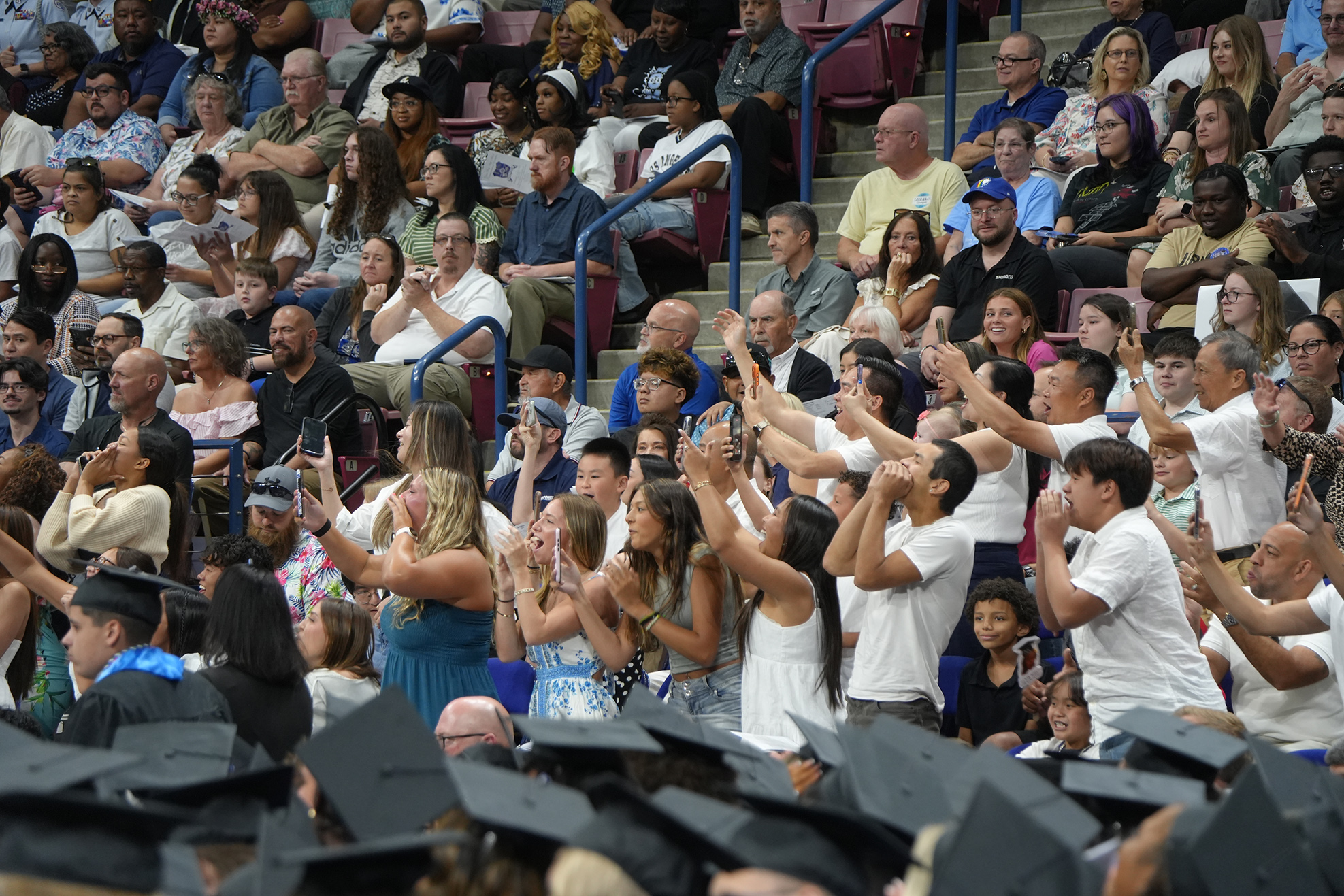 A crowd in a stadium with many people clapping, some wearing graduation gowns and hats.