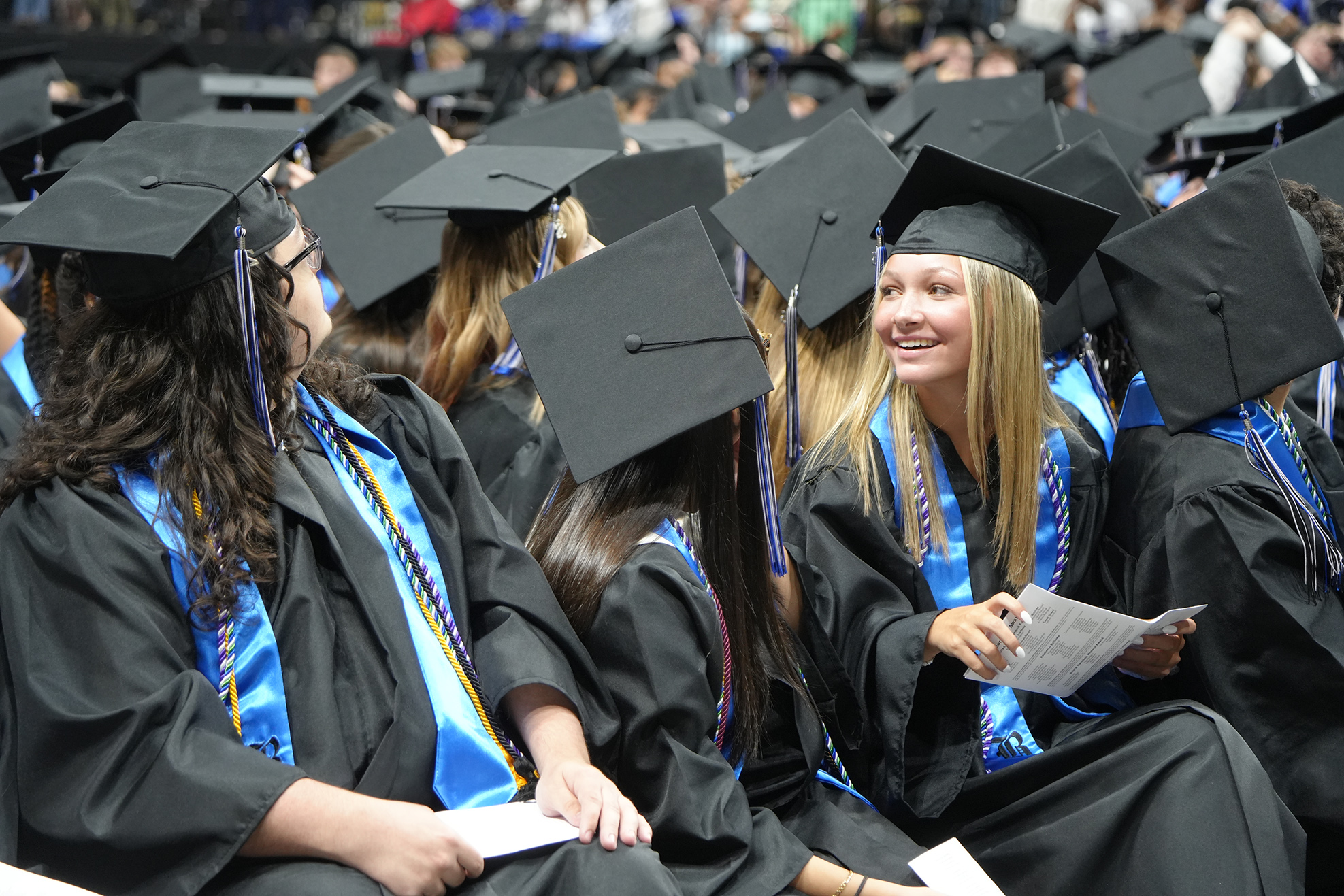 Graduates in black caps and gowns sit in rows at a ceremony. Some hold papers, and many face forward.