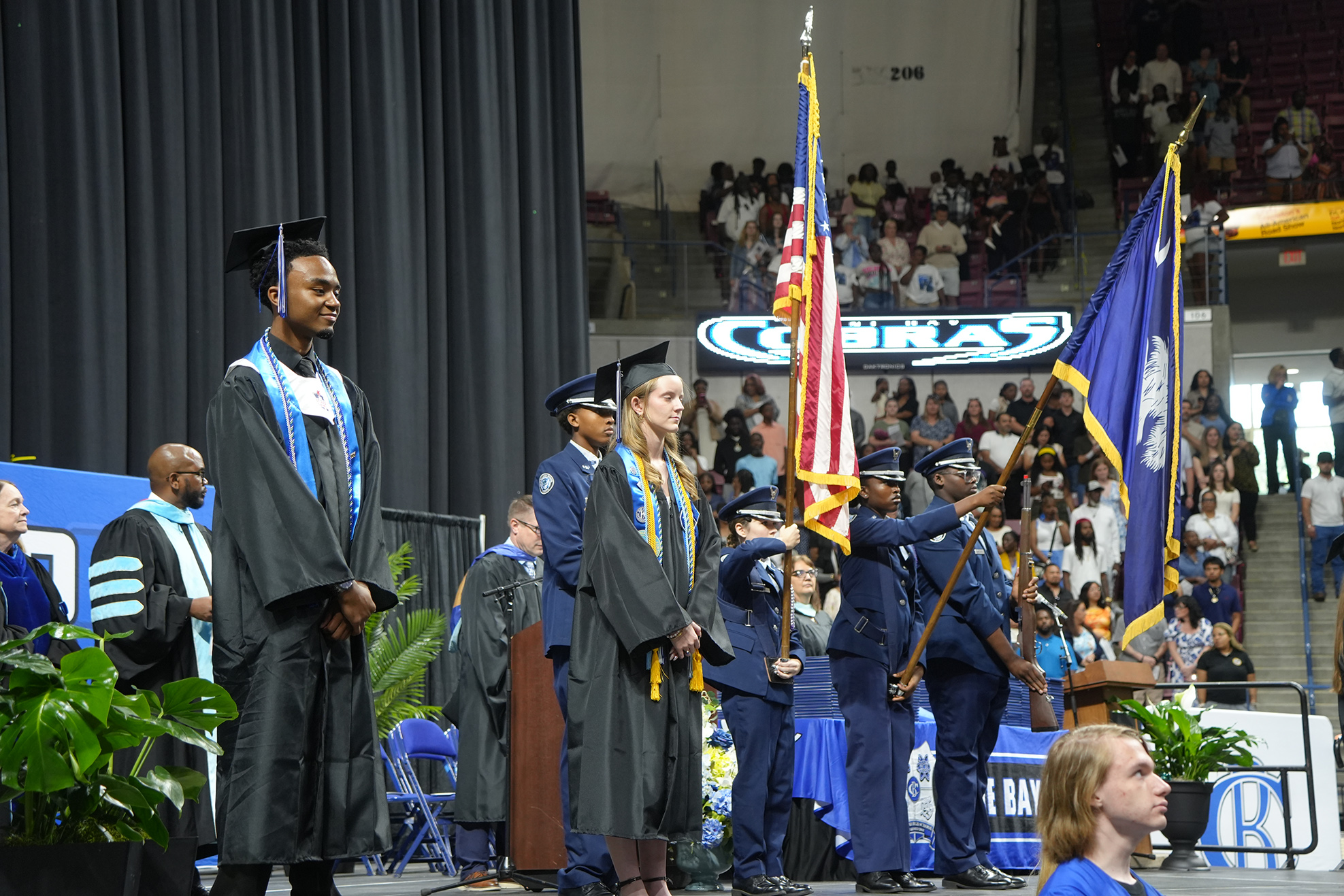 Graduates in caps and gowns stand on a stage with American and school flags held by uniformed officers.