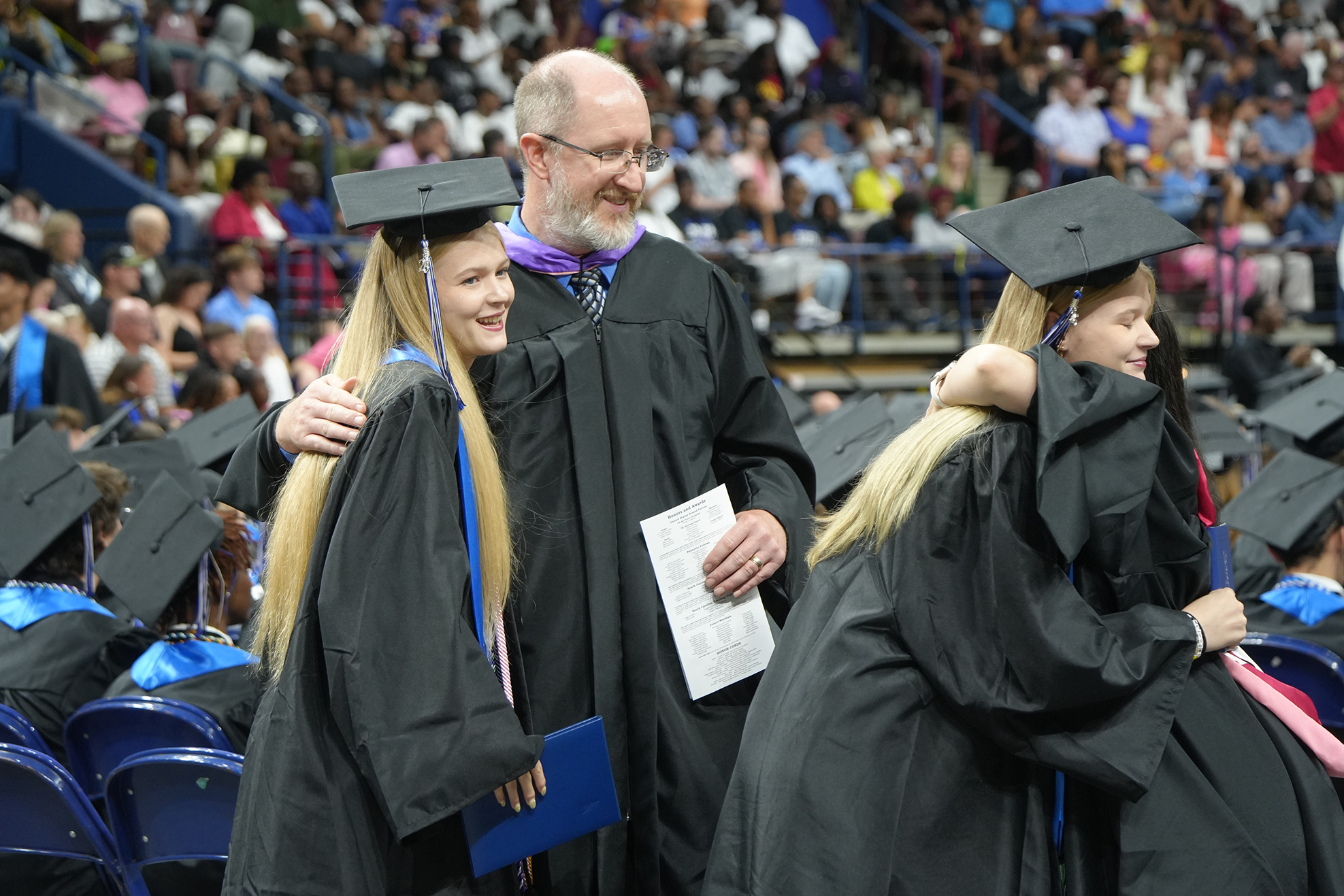 Graduates in caps and gowns stand in a stadium, one holding a paper, with a crowd in the background.