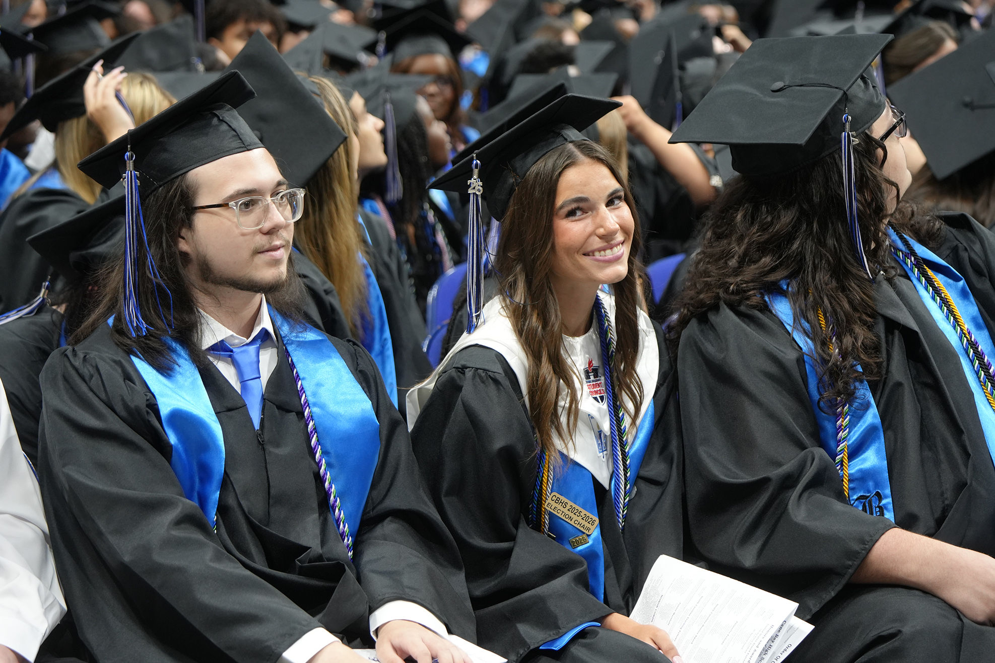 People in graduation gowns and caps, seated in a row, with some smiling. The background is blurred.