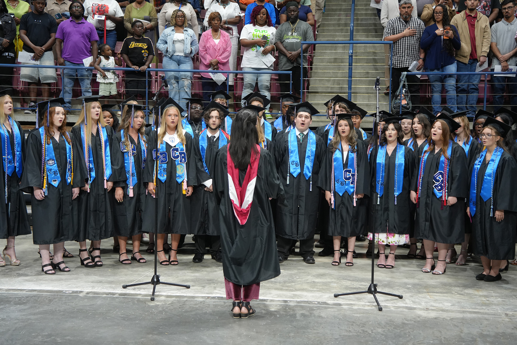 A group of students in graduation gowns performs on stage with a crowd of people watching.