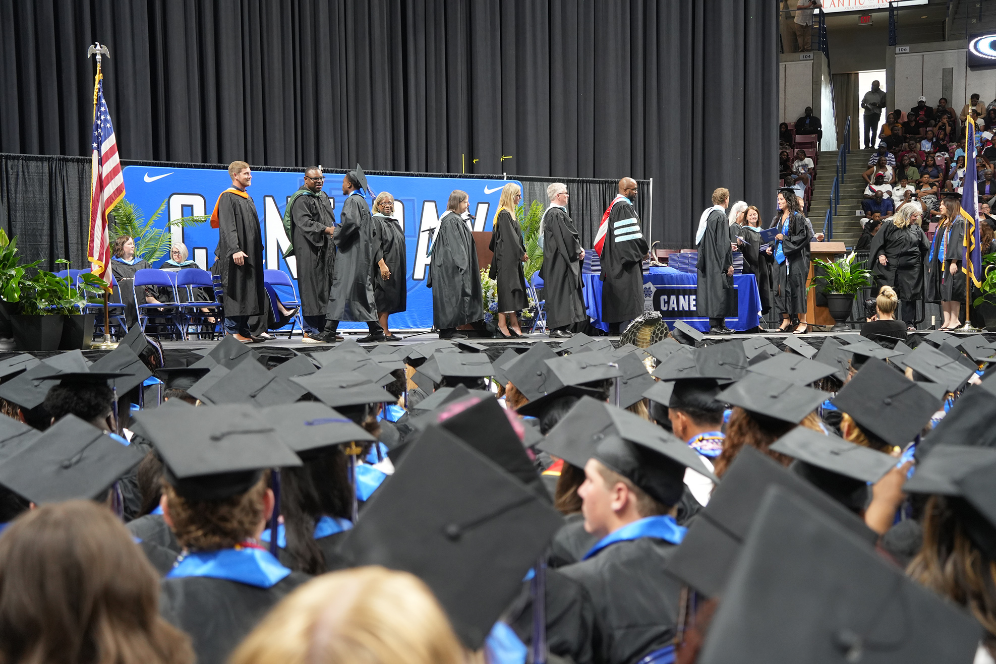 A graduation ceremony with graduates in gowns and caps, standing on stage. The audience sits below in front of a stage backdrop.