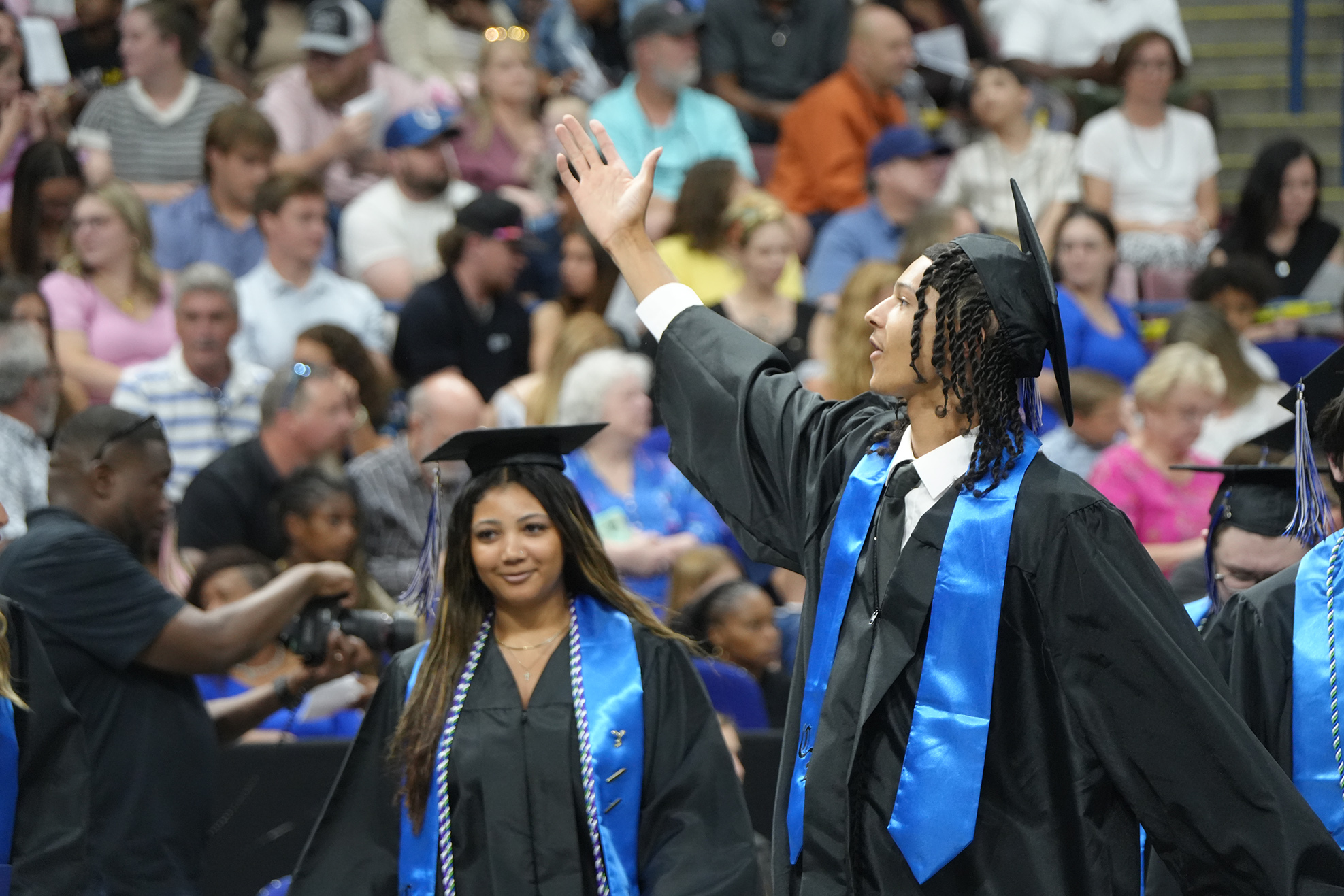 Graduates in black gowns and caps walk in front of a seated audience at a graduation ceremony.