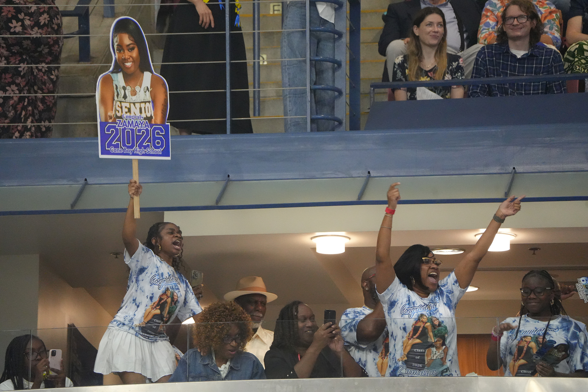 Spectators in a stadium stand with raised arms, cheering. Two individuals in blue shirts with a woman's photo hold up a sign reading "2026".