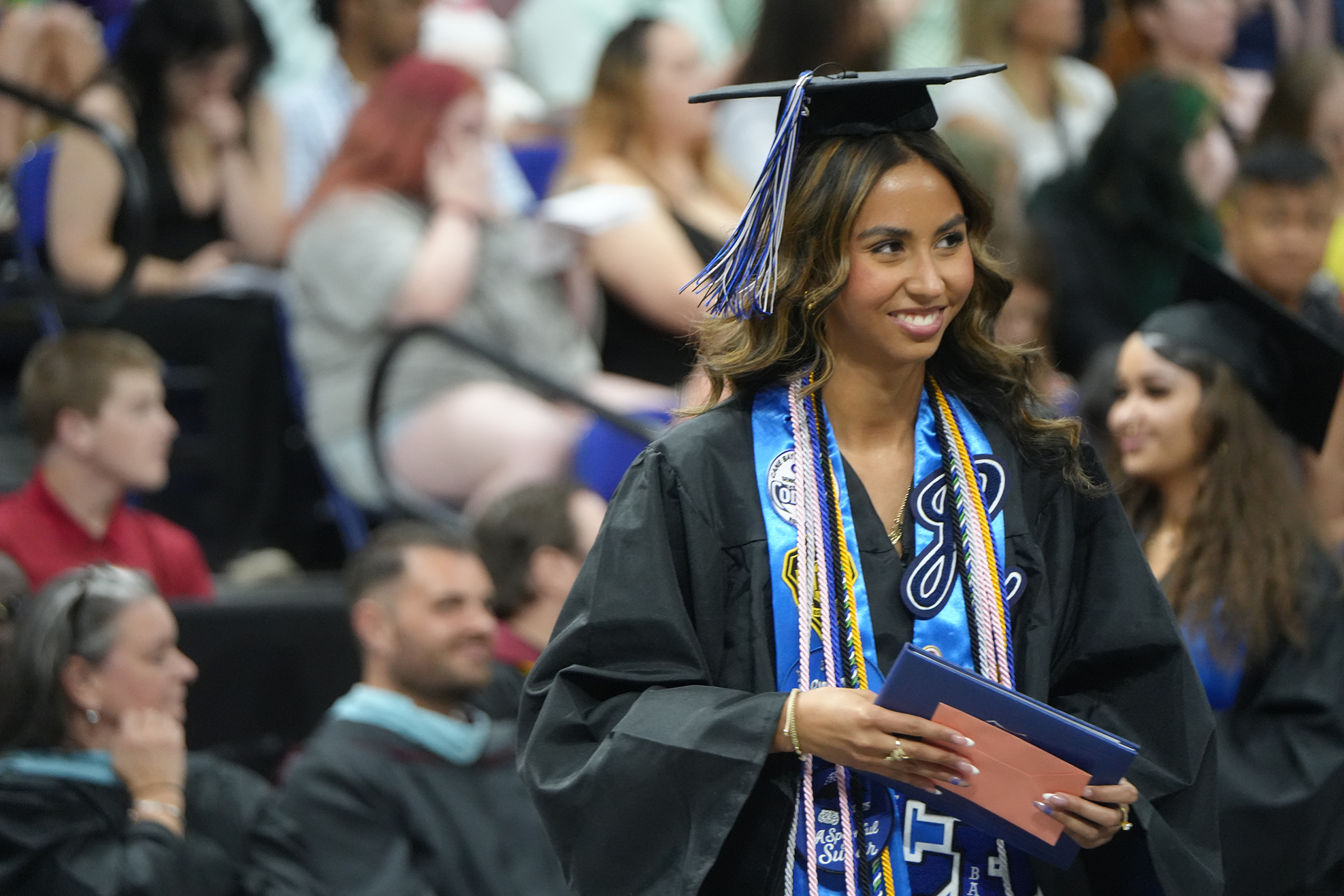 A smiling woman in a black graduation gown with blue and white sashes walks in an audience.