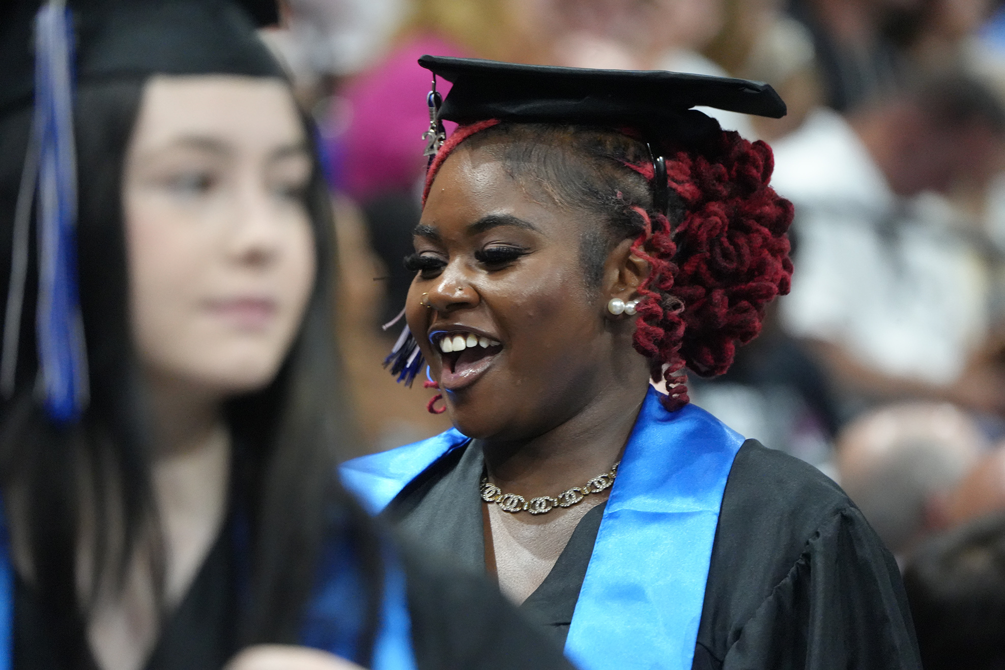 Two women in graduation gowns, one with a blue stole, smiling at a ceremony; blurred crowd in background.