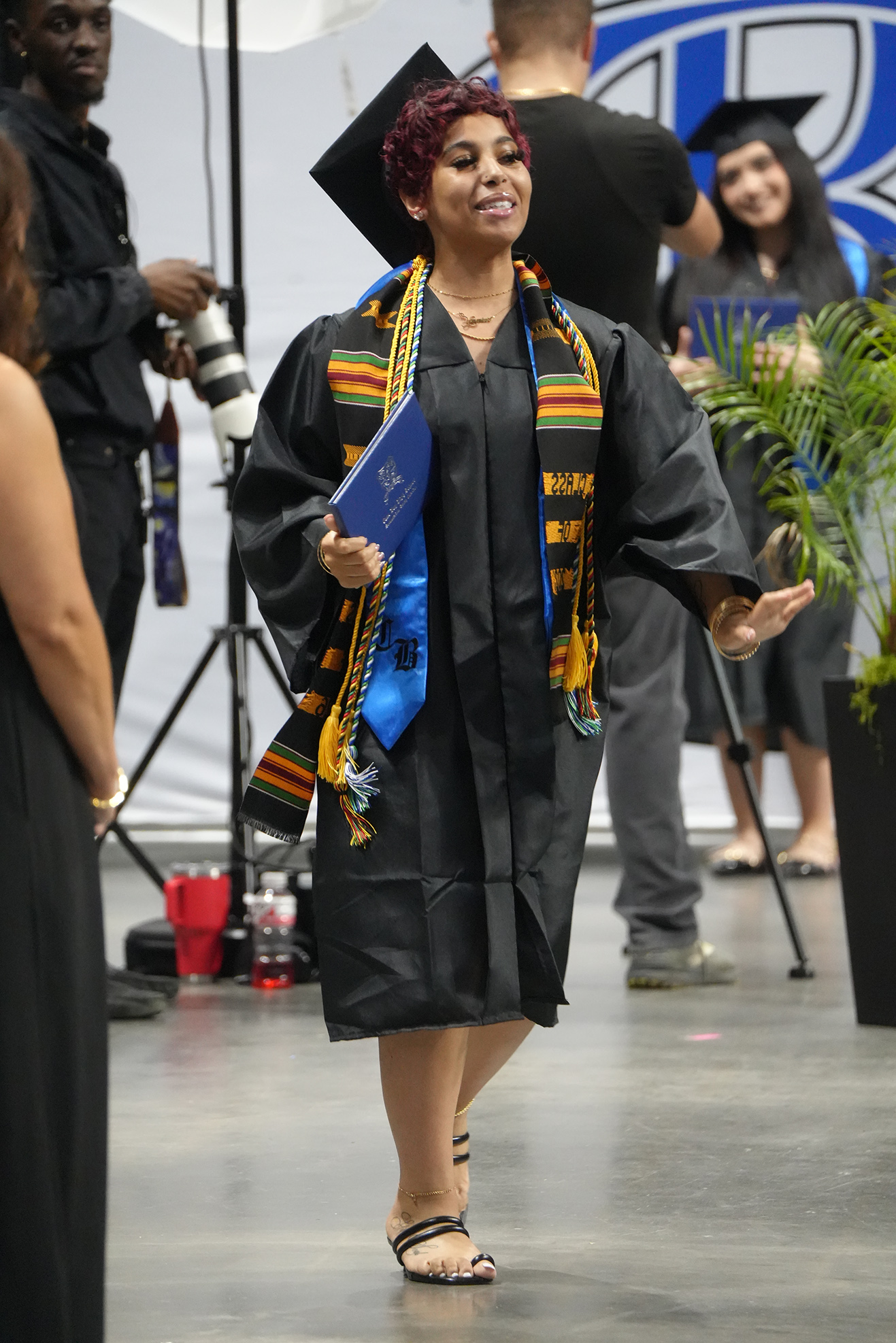 A woman in a graduation gown walks on stage while holding a blue folder. A green plant and a tripod stand behind her.