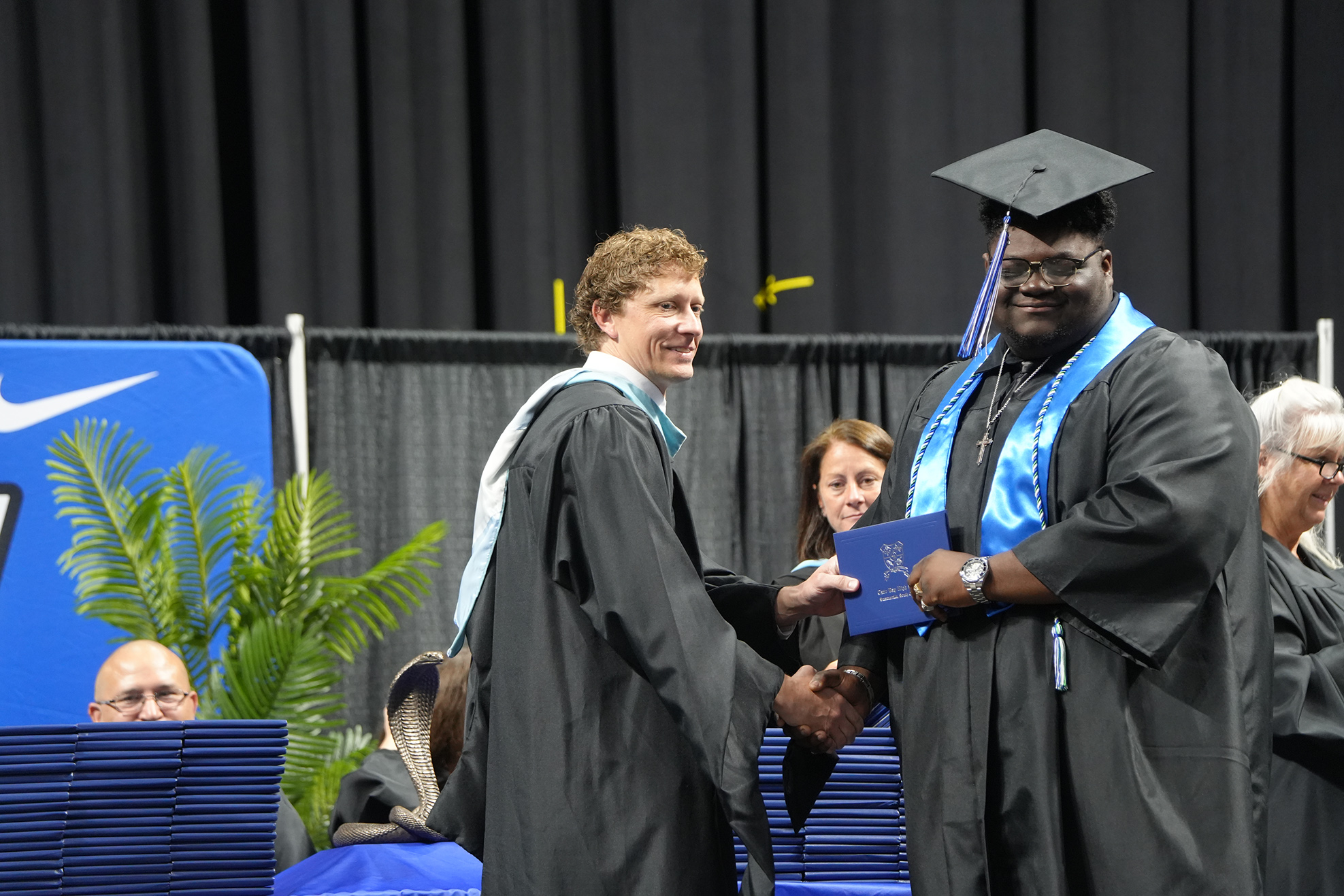 A male graduate in a cap and gown shakes hands with a faculty member during a ceremony.