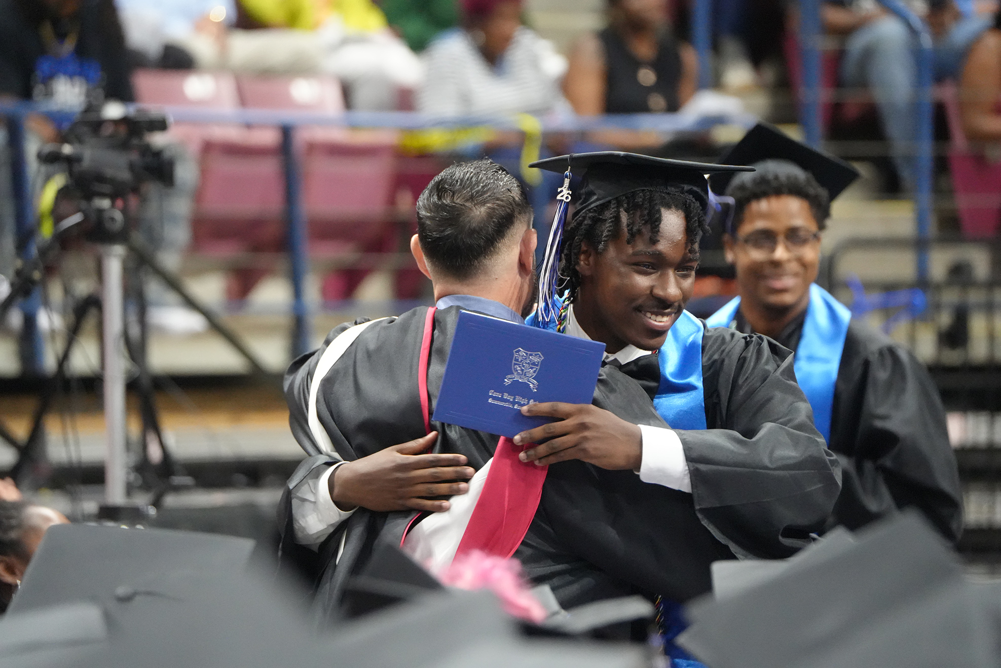 Graduates in black gowns, one holding a blue book, embrace each other. Behind them, rows of spectators in a stadium.