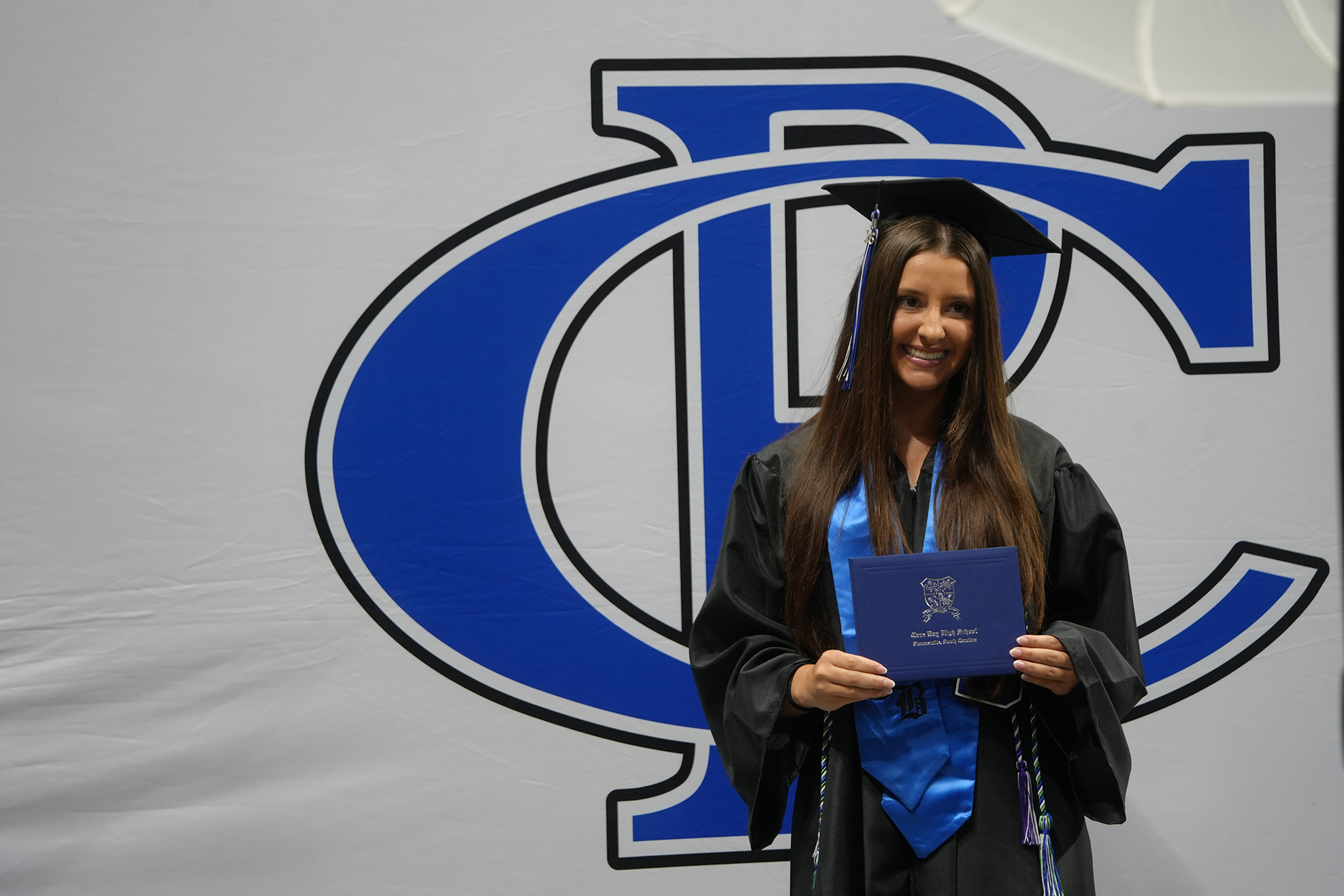 Woman in cap and gown holding diploma, standing in front of a blue backdrop with a large letter D.