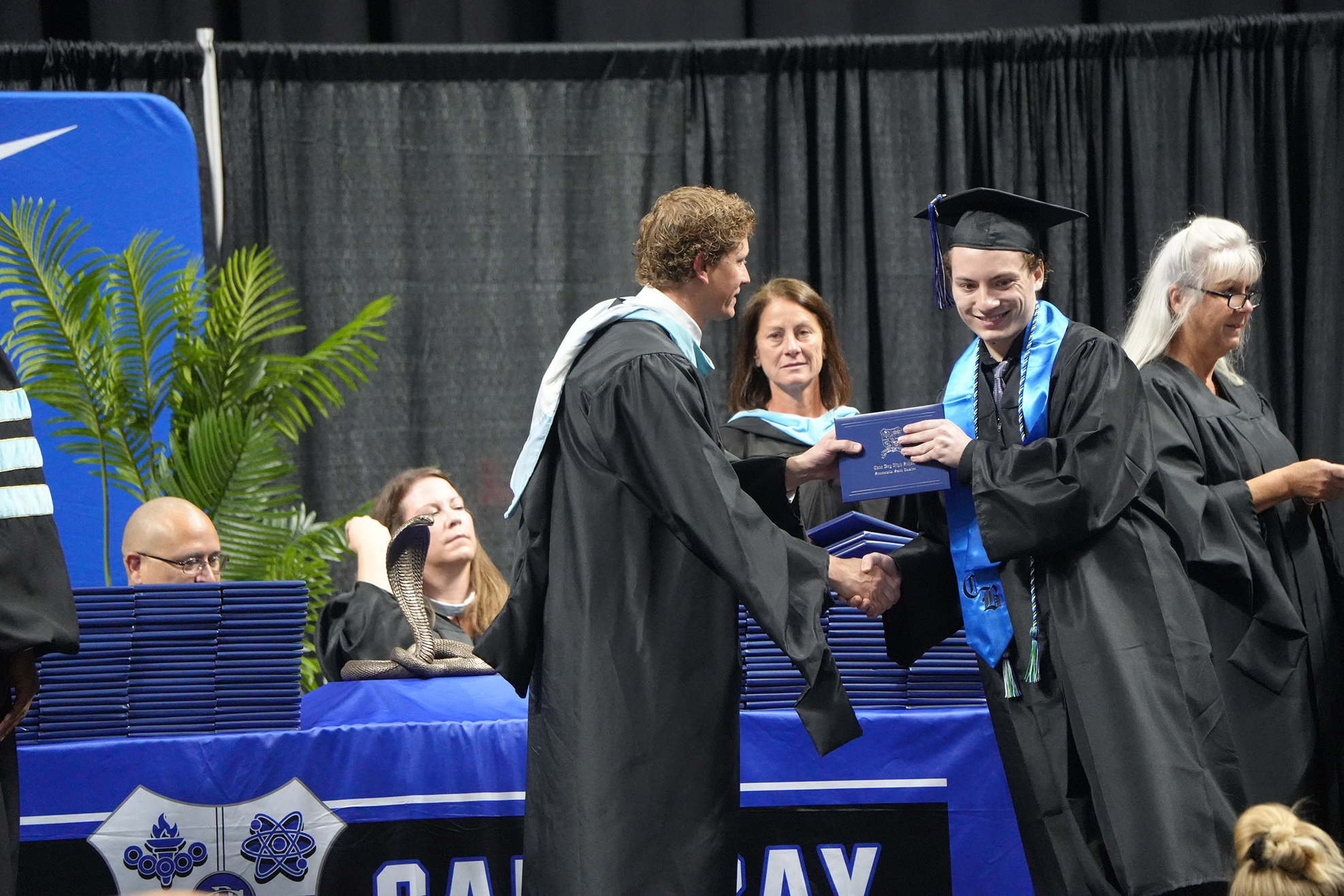 A man hands a diploma to another man at a graduation ceremony; people sit behind them.