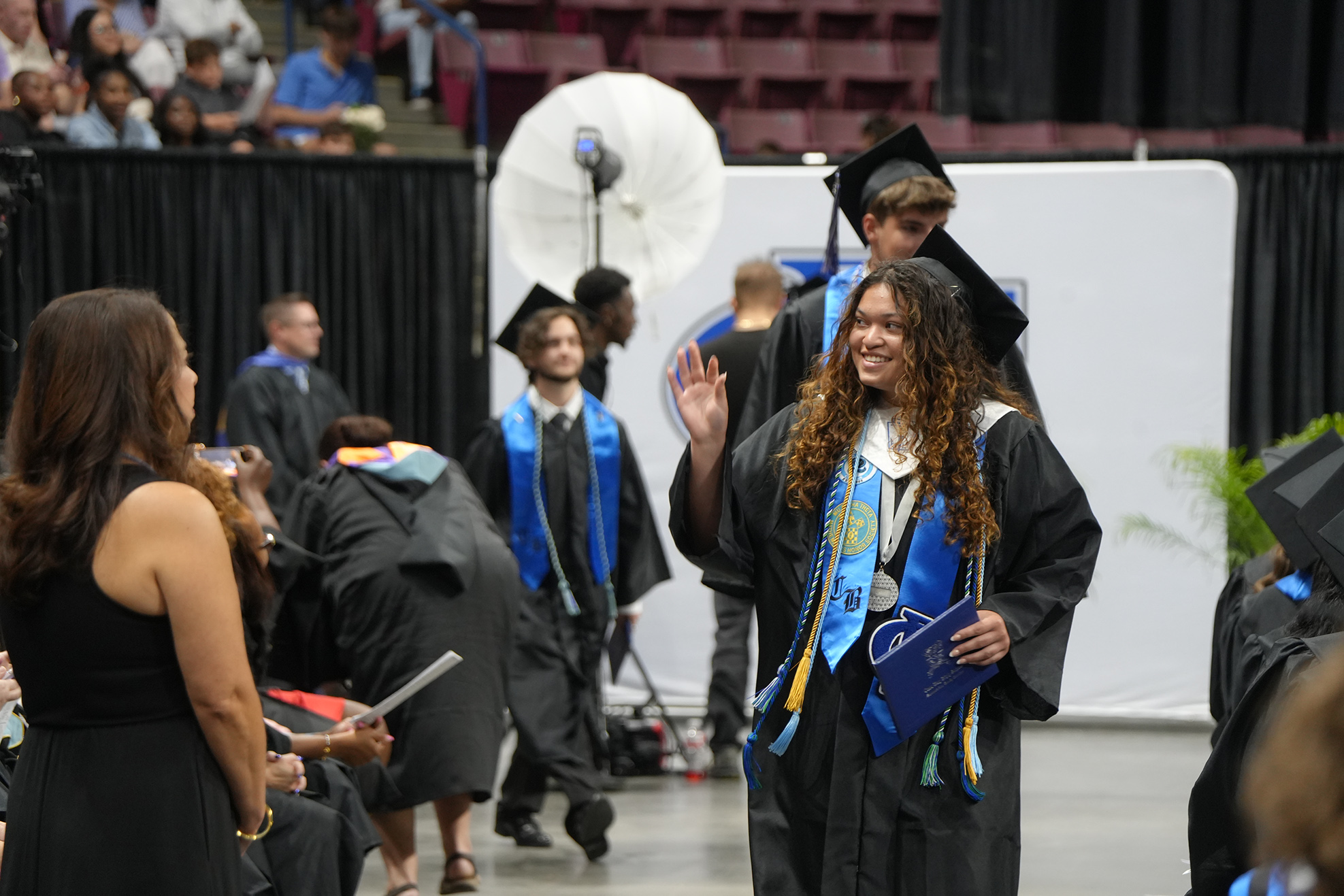 A group of people wearing academic regalia are standing in a hallway. One woman holds a folder and smiles.