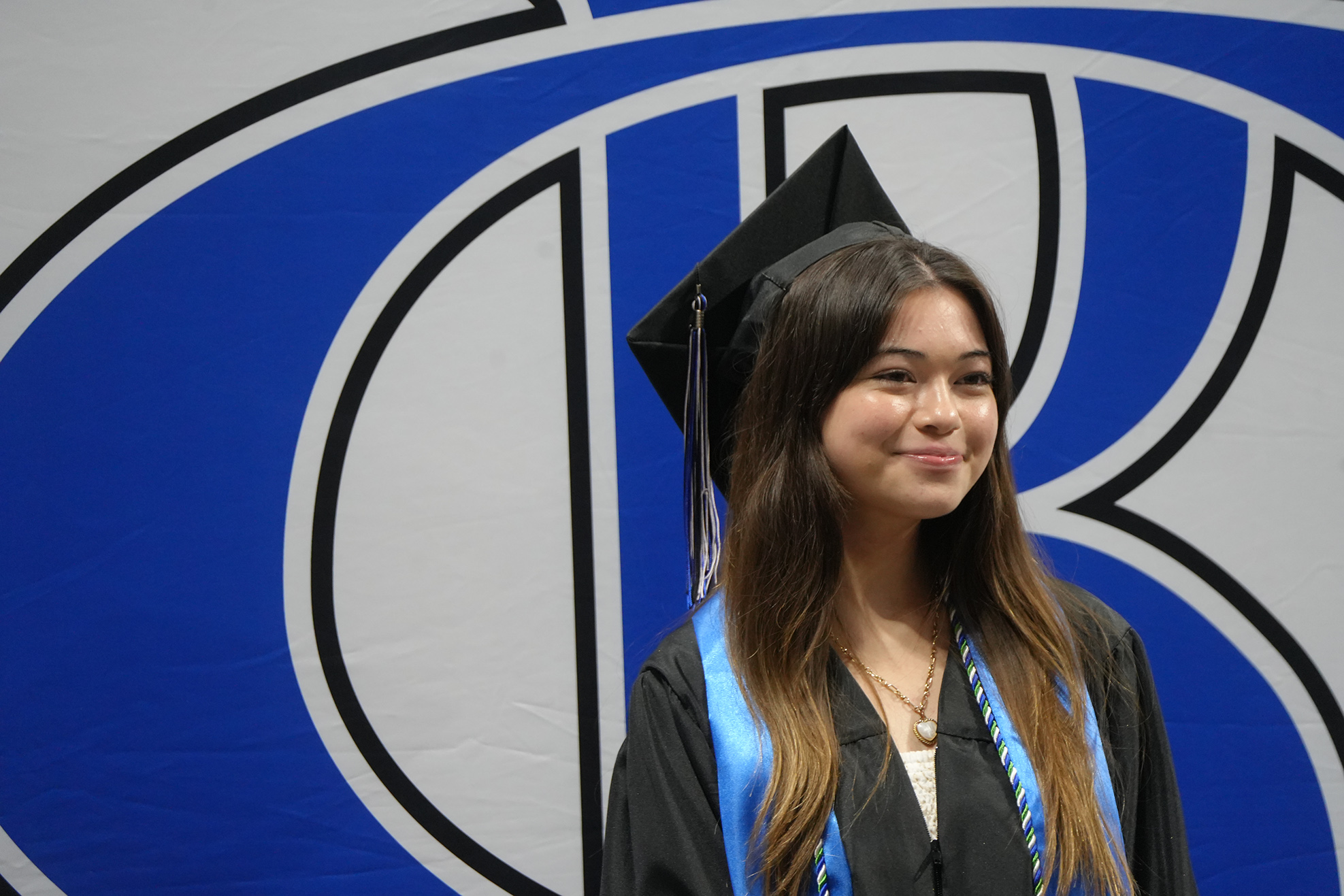 A woman in a black graduation gown and cap stands before a blue and white backdrop.