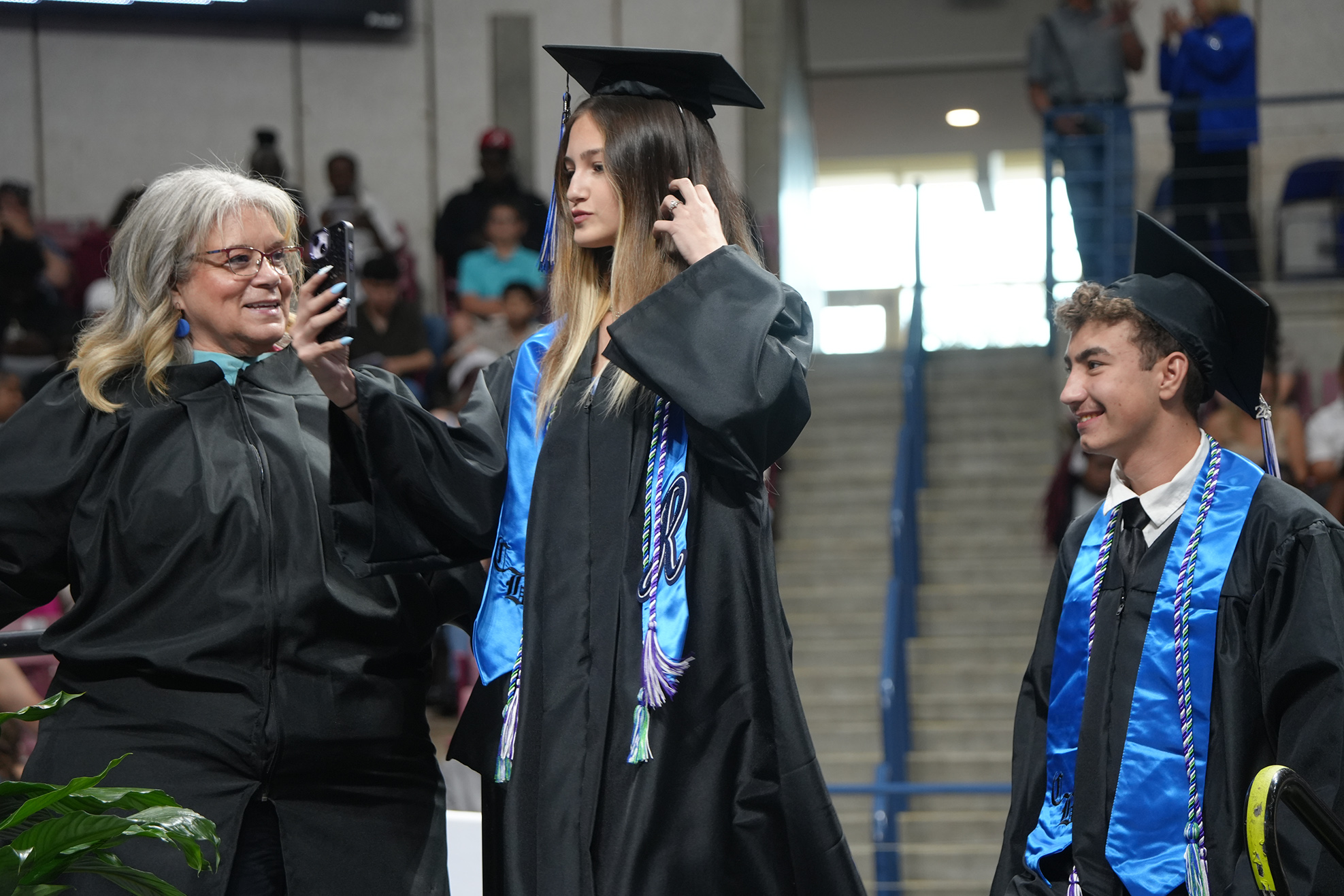 A woman adjusts her graduation cap while standing with two others, one of them holding a phone.