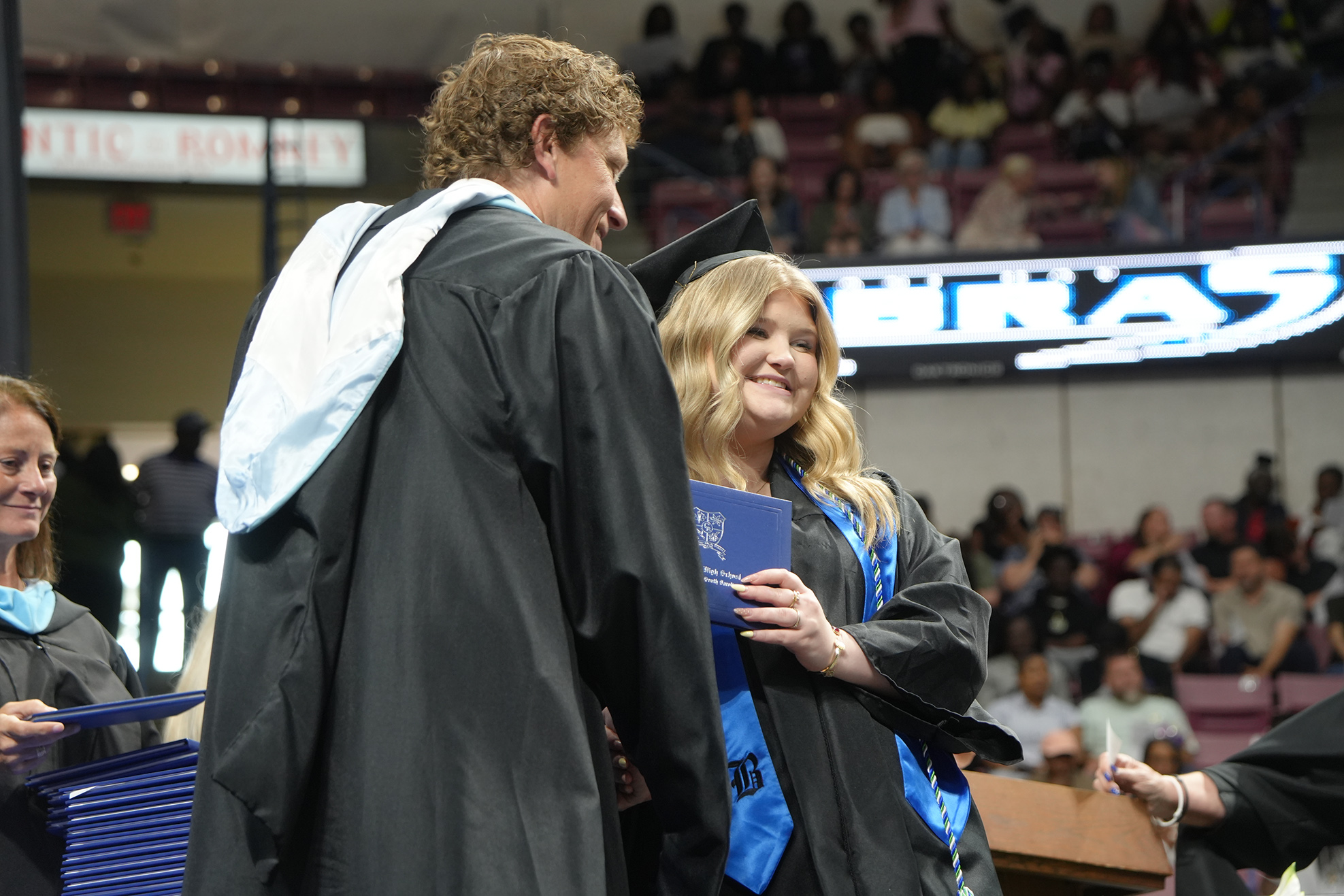 Two individuals in academic regalia are facing each other. One holds a diploma, and the other holds a folder.